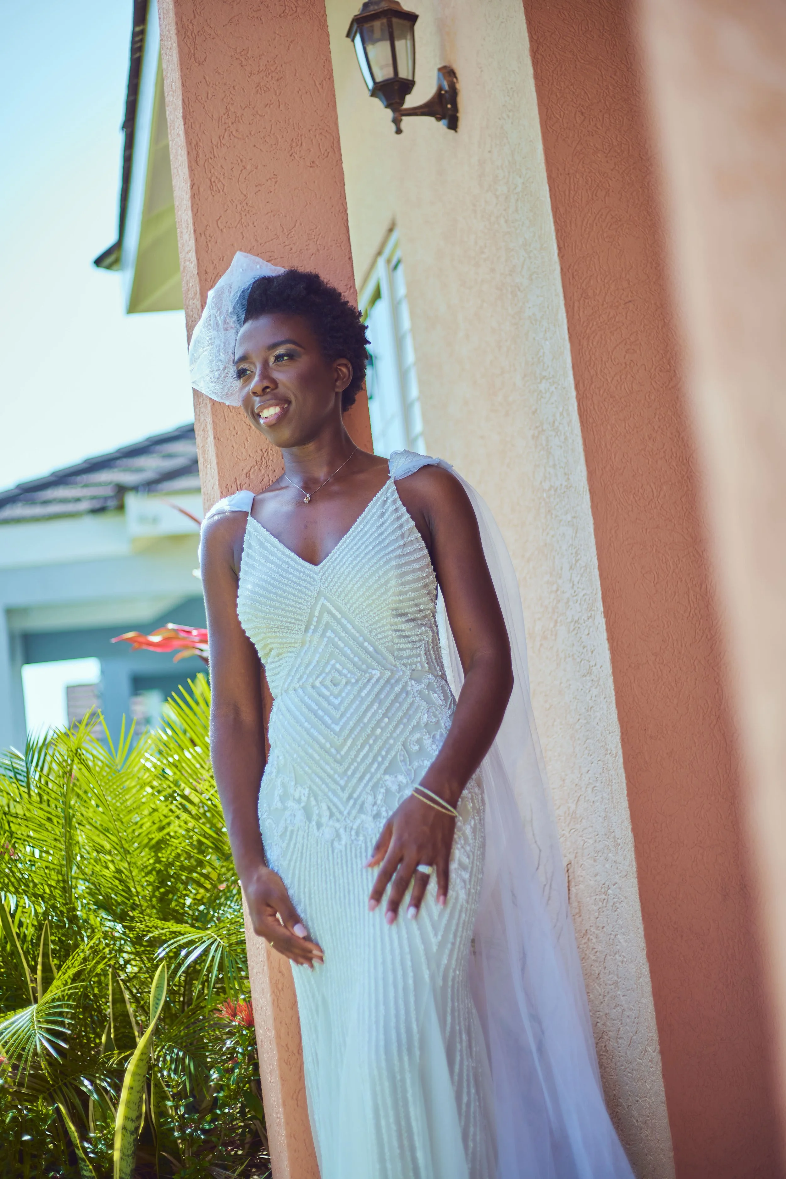 A woman in a white wedding dress standing outside against a peach-colored wall, smiling and looking away. She has short curly hair, a veil, and is adorned with jewelry, with greenery visible in the background.