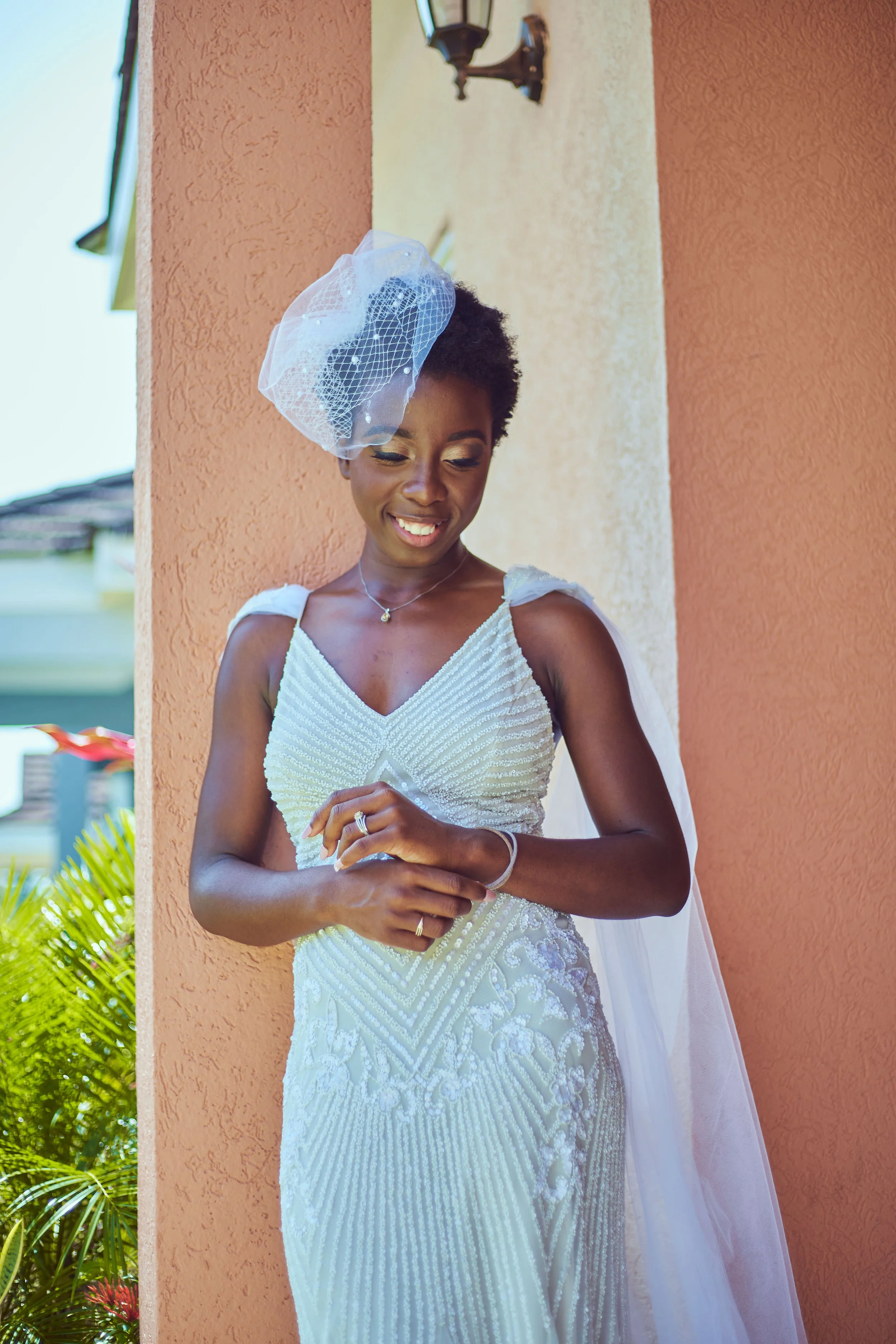 A bride in a white wedding dress standing outdoors beside a peach-colored wall, smiling and looking down, with a veil on her head.