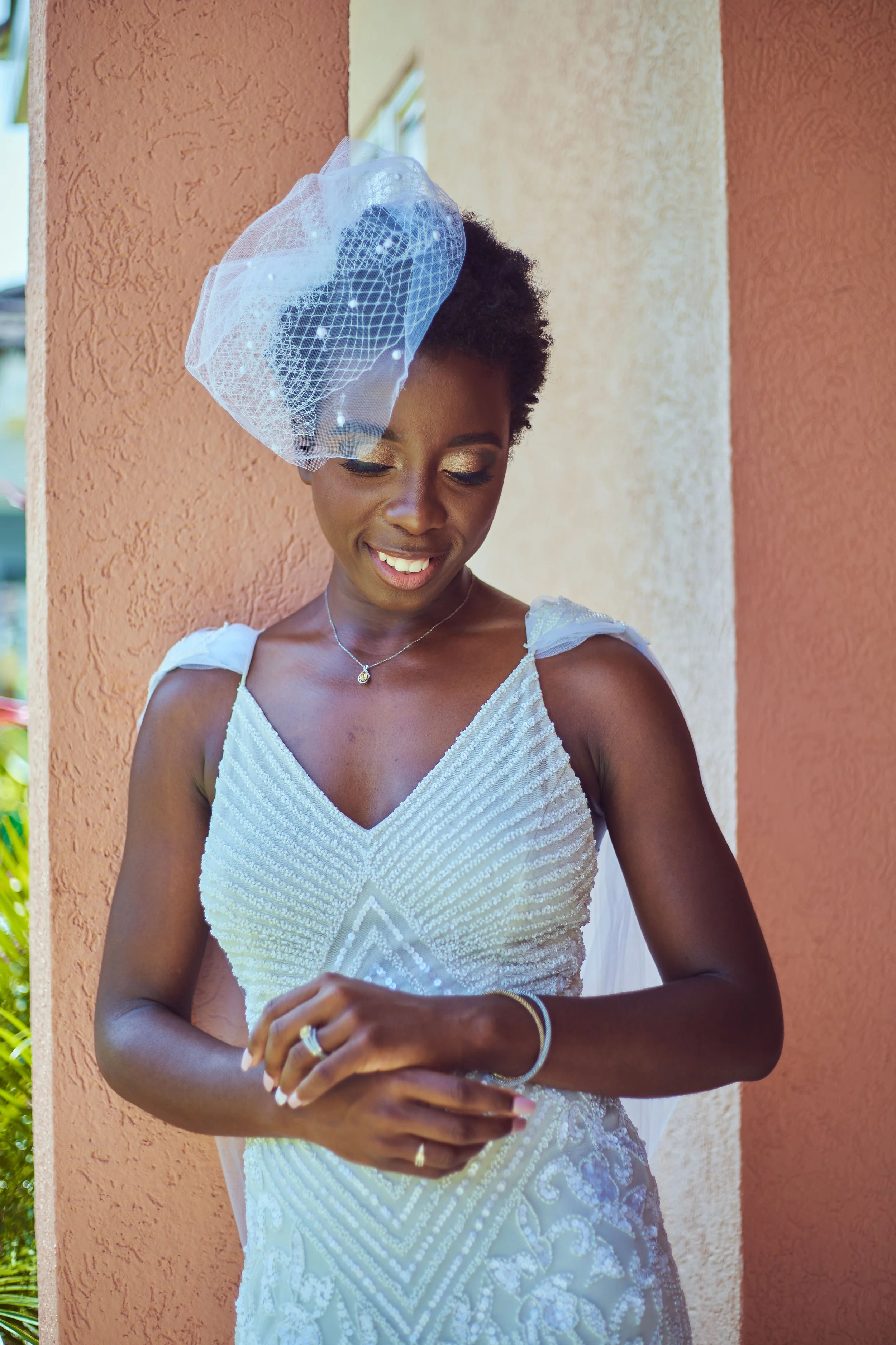 A woman in a white wedding dress standing outdoors, smiling, with a pink wall and some greenery in the background.