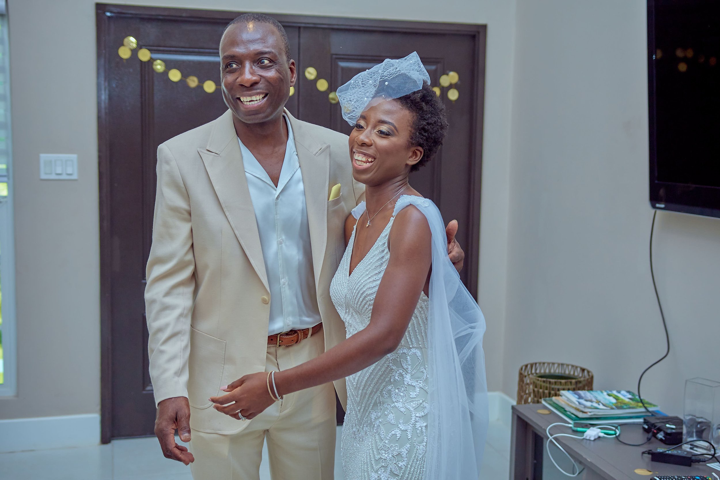 A man and a woman dressed in formal attire, smiling and holding hands, standing indoors in front of a dark wooden door. The woman is wearing a white dress with intricate lace patterns and a tulle veil, while the man is in a light-colored suit with a 