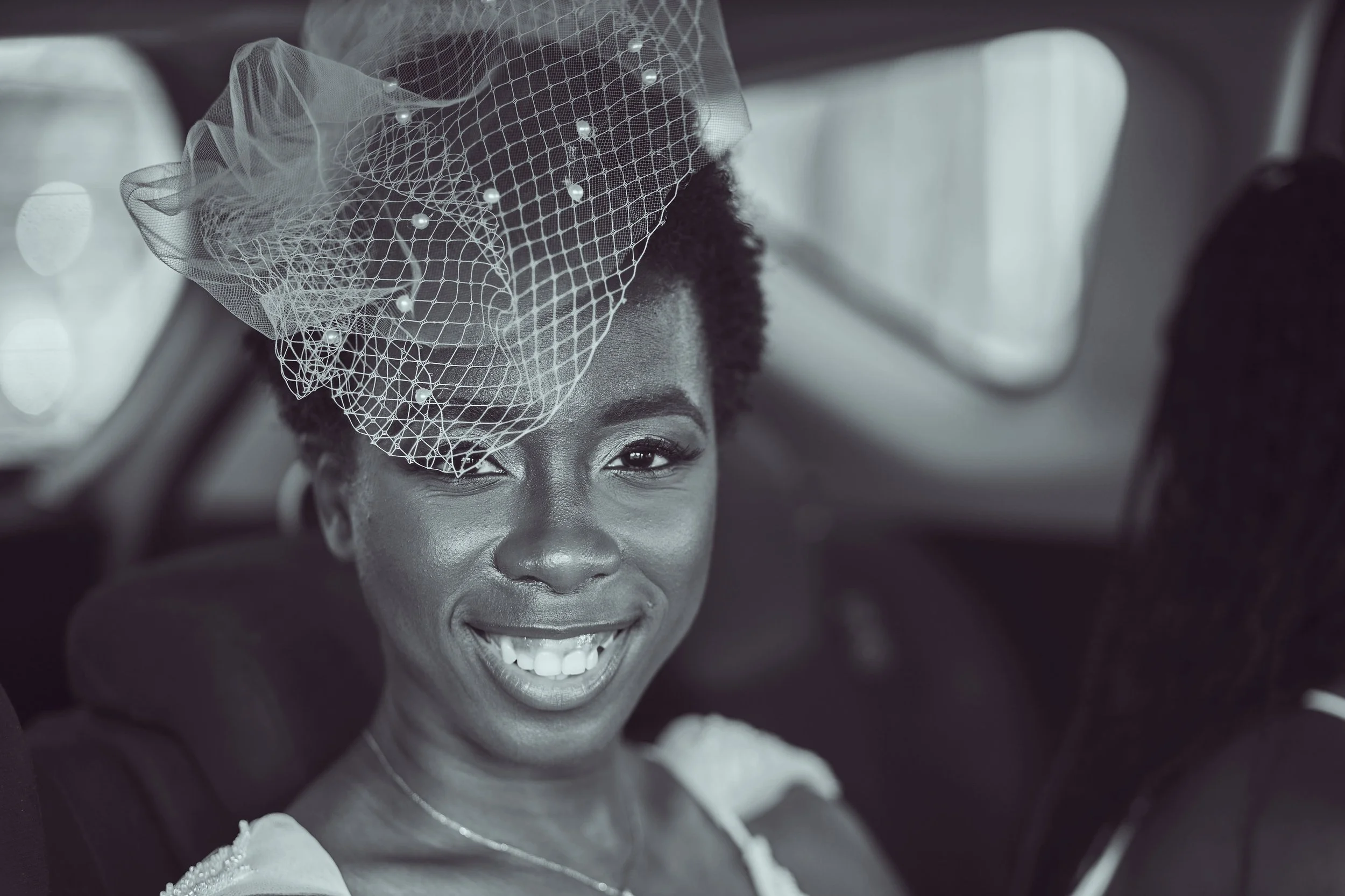 A smiling woman with short curly hair wearing a bridal veil, sitting inside a vehicle.