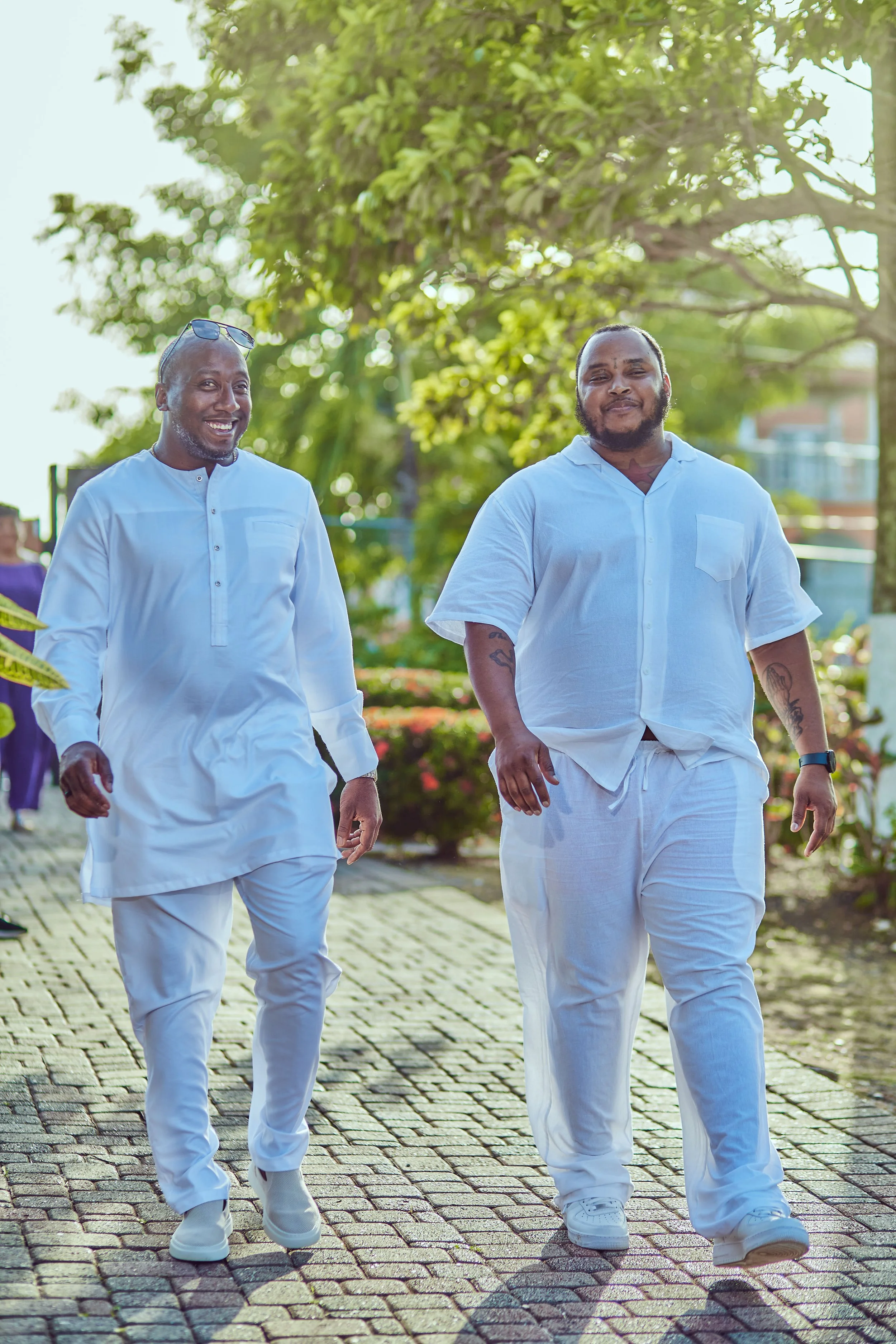 Two men wearing white clothing walking outside on a sunny day, smiling, with trees and greenery in the background.
