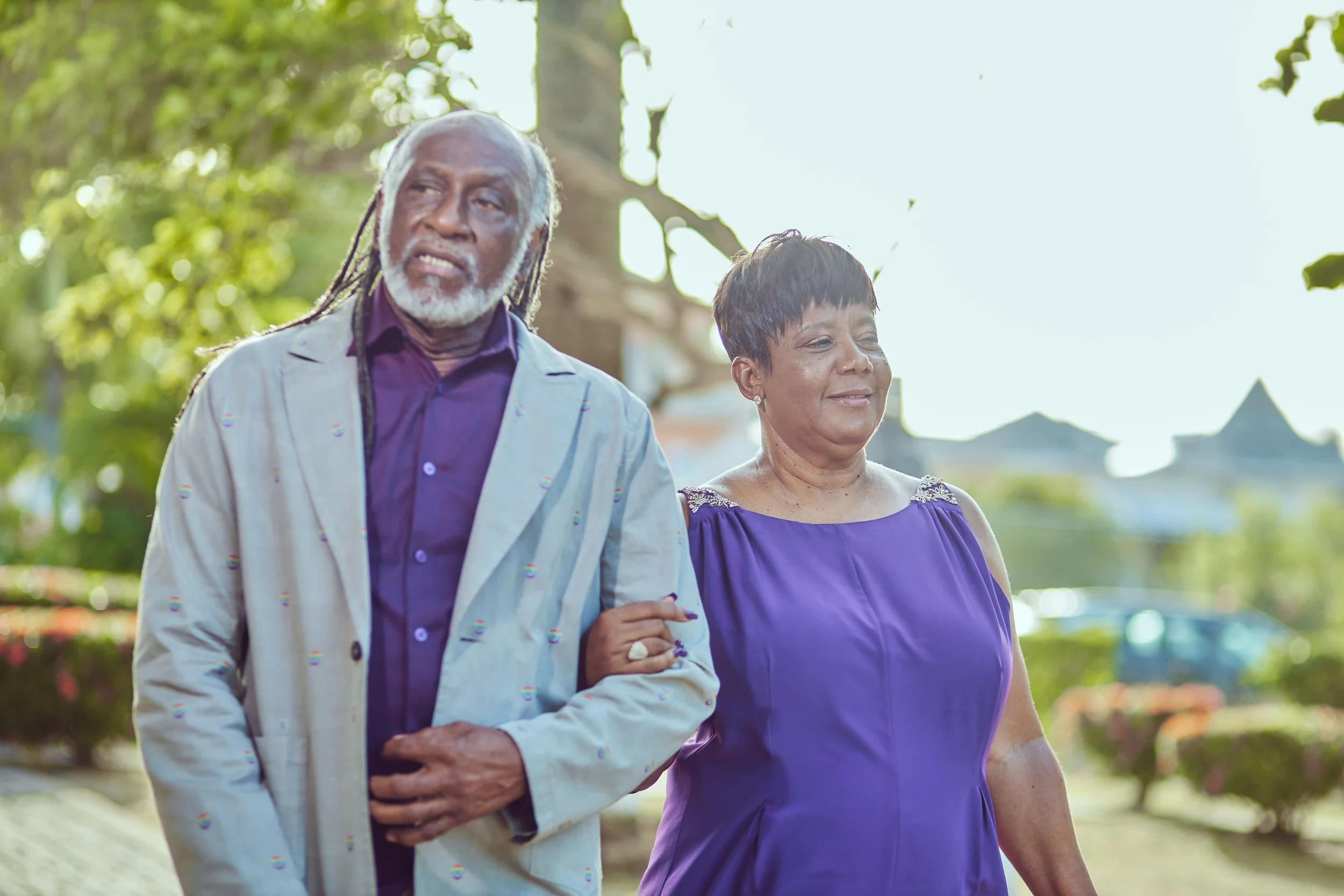 An elderly couple walking outdoors in a park, holding hands. The man has long gray hair and a beard, wearing a light-colored jacket over a dark shirt. The woman has short dark hair, wearing a purple dress with embellished shoulders, and is smiling.