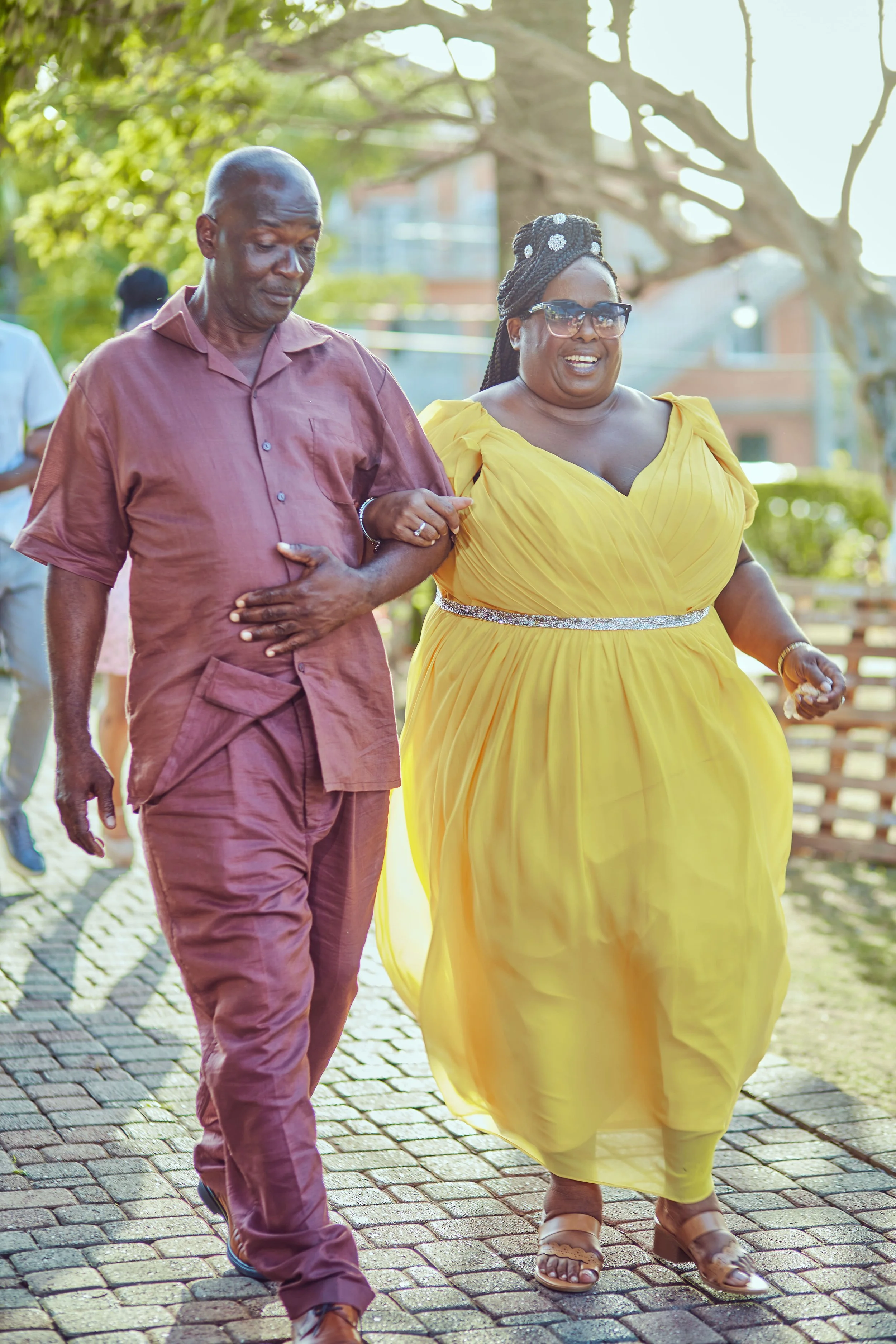 A man and woman walking outdoors on a sunny day, holding onto each other's arms, with trees and a building in the background. The woman is wearing a bright yellow dress and glasses, and the man is in a maroon shirt and pants.