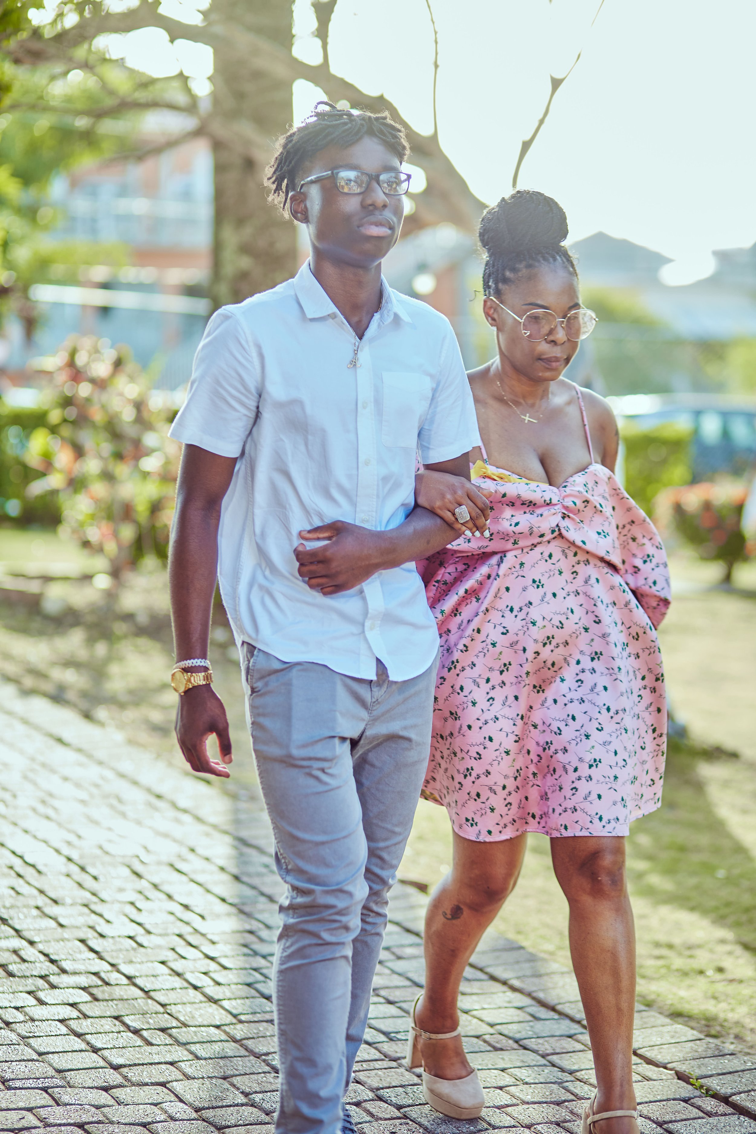 A woman with glasses wearing a pink floral dress is being assisted by a young man with glasses, wearing a white shirt and grey pants, as they walk outside on a sunny day.