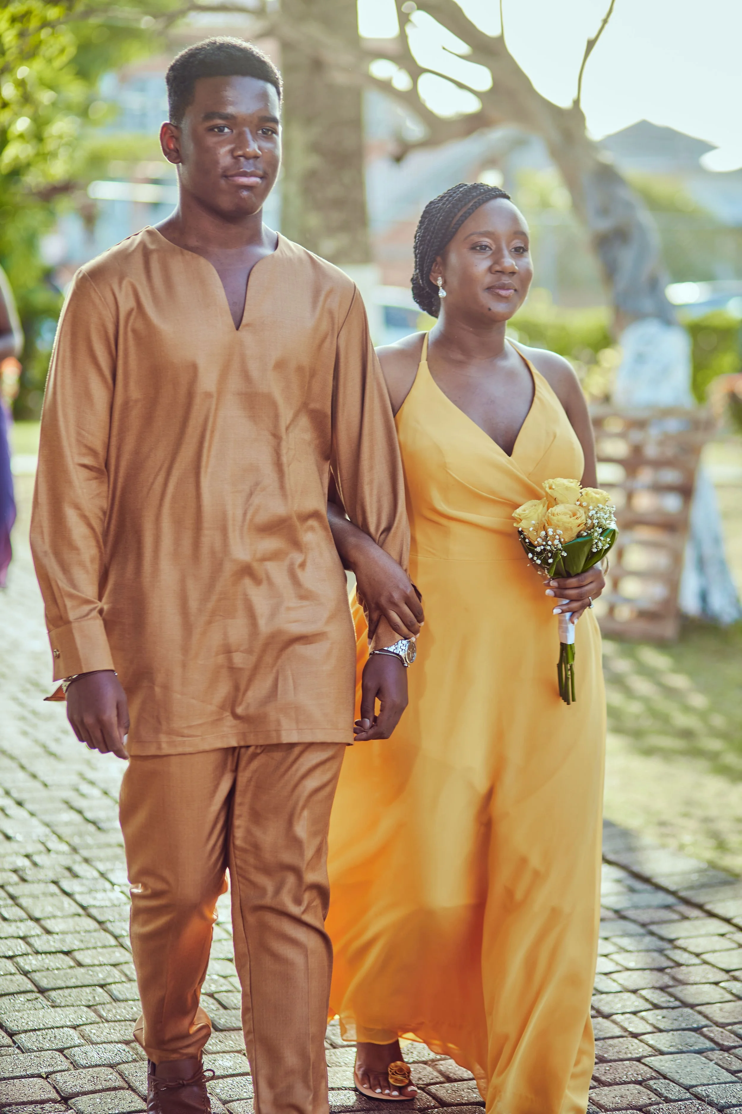 A young man in a gold traditional outfit walking arm-in-arm with a woman in a yellow dress holding a bouquet of yellow flowers, outdoors during daytime.