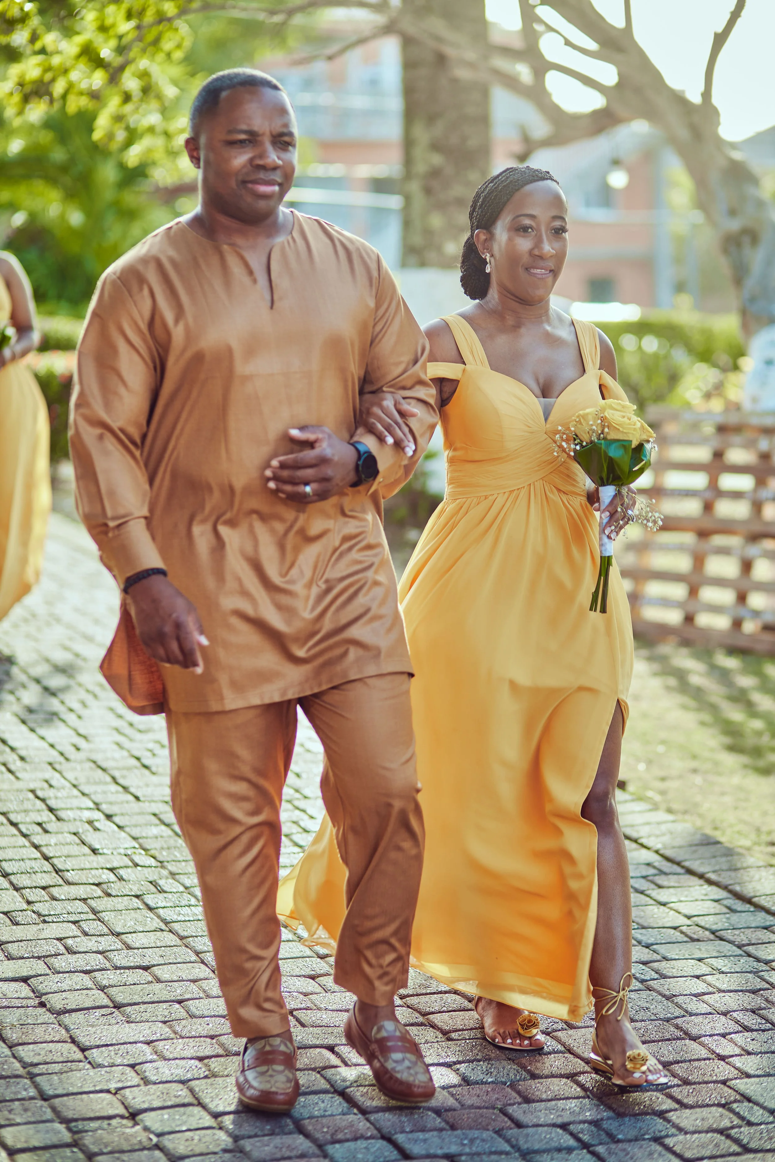A couple dressed in matching gold outfits walking on a paved walkway outdoors, with the woman holding a bouquet of flowers.