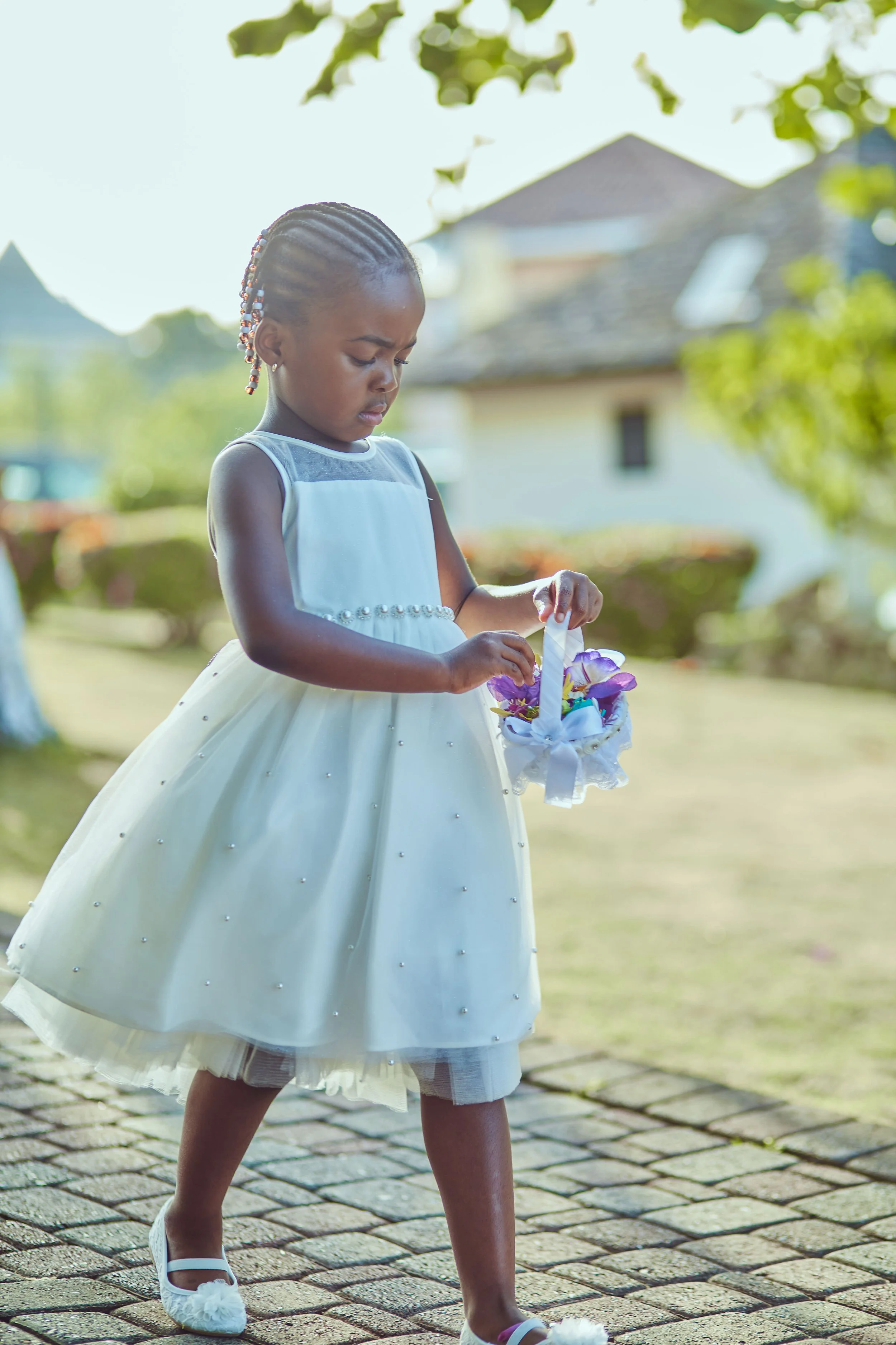 A young girl in a white dress with pearl detail holding a basket of purple and white flowers outdoors.