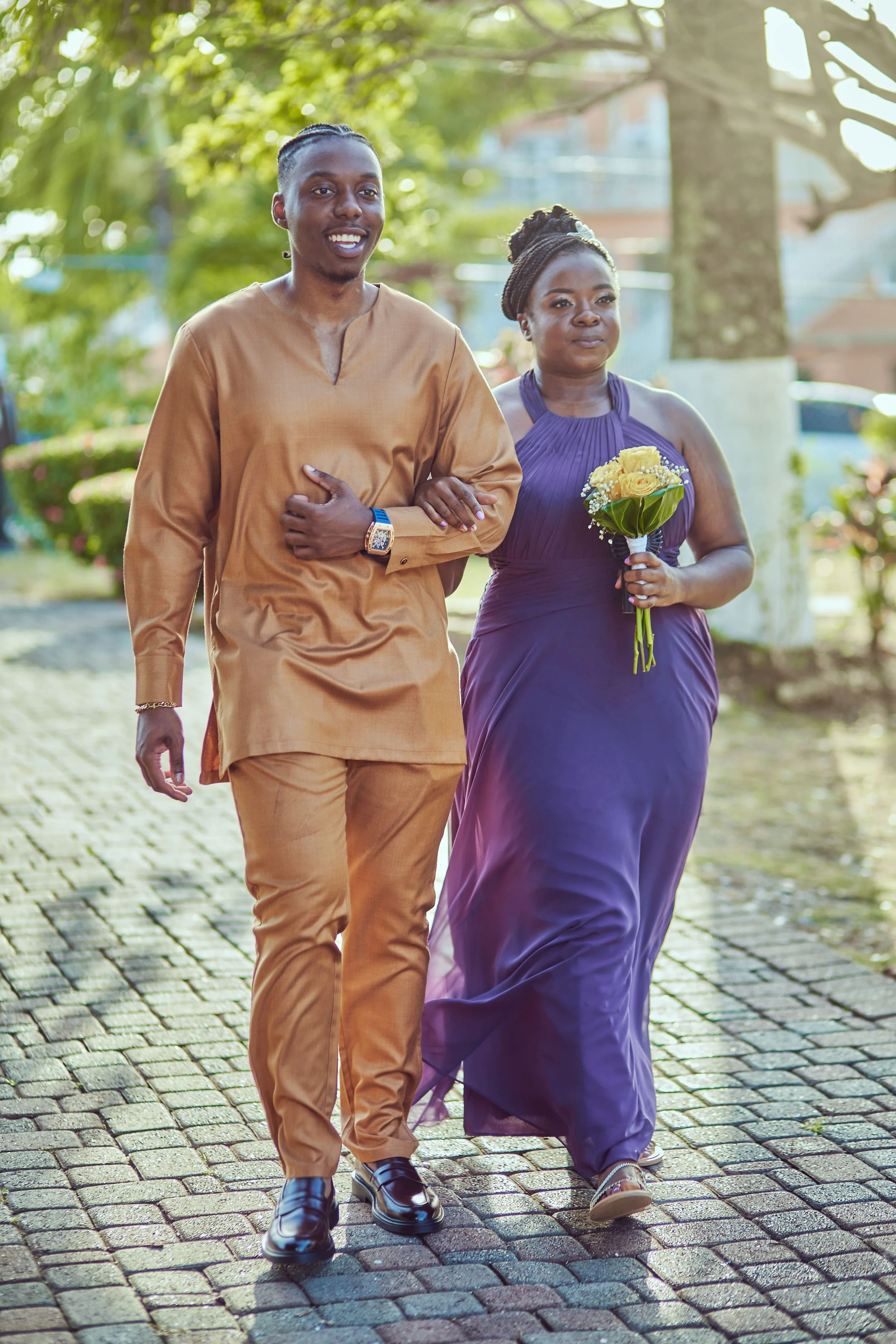 A man in a light brown traditional outfit and a woman in a purple dress walking arm-in-arm outdoors. The woman holds a bouquet of yellow flowers. They are on a cobblestone path surrounded by greenery, with sunlight filtering through the trees.