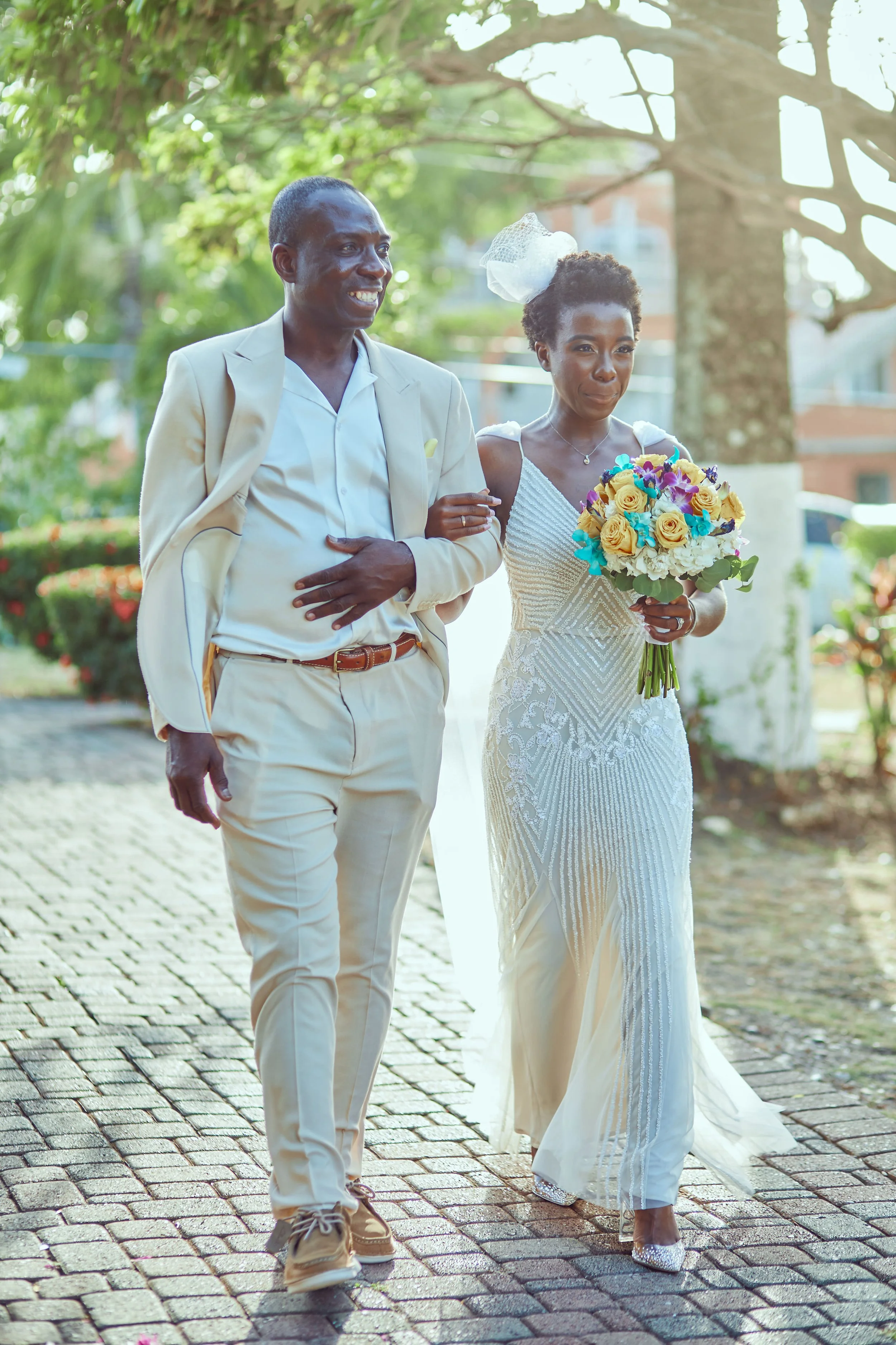 A woman in a white wedding dress holding a bouquet of yellow, purple, and white flowers, walking arm-in-arm with a man in a light-colored suit on a cobblestone path outdoors, with trees and sunlight in the background.