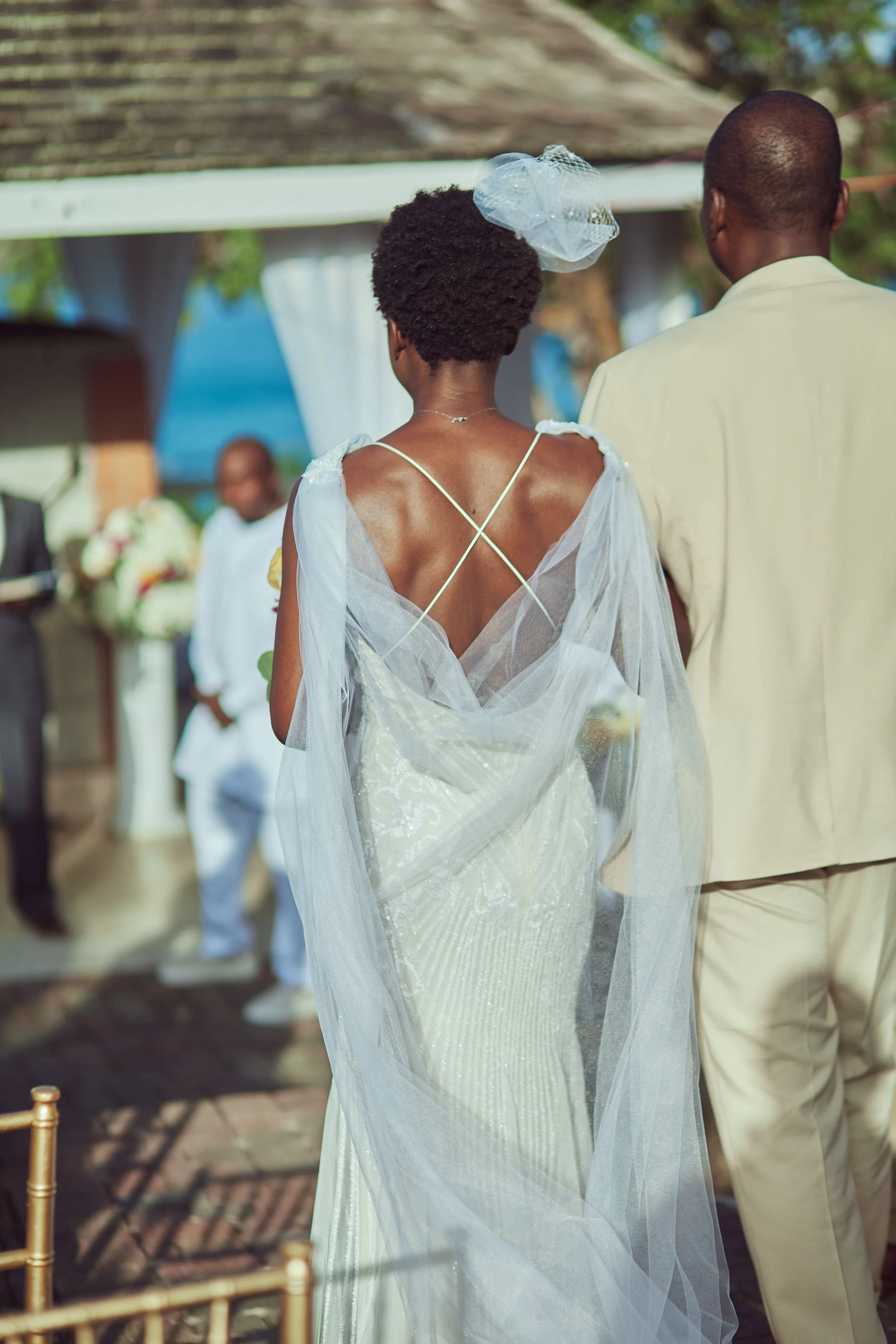 A bride and groom at an outdoor wedding ceremony, viewed from behind. The bride is wearing a white dress with a tulle veil, and the groom is in a cream-colored suit. There are guests and floral arrangements in the background.
