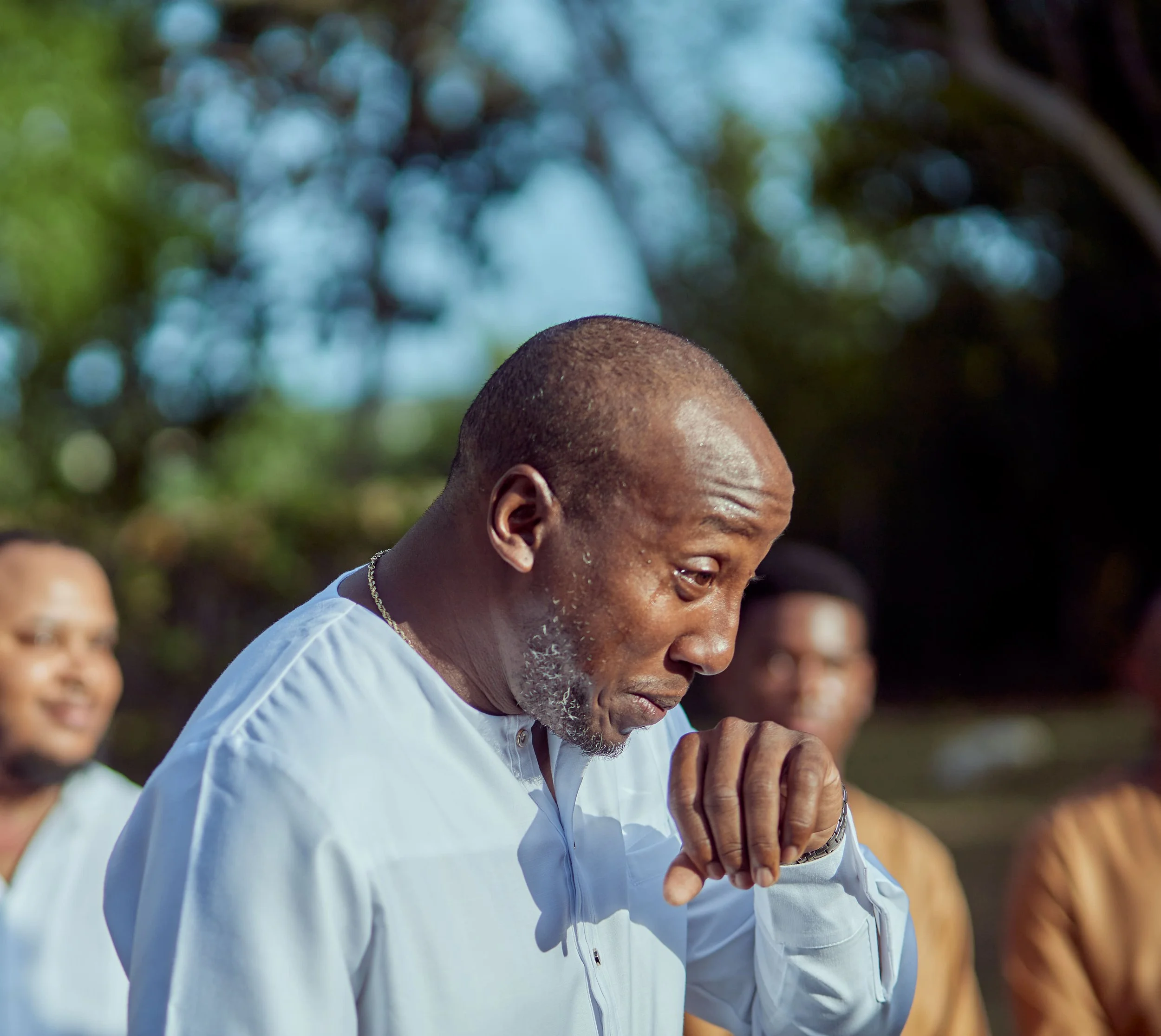A man in a white shirt with short hair and a beard, appearing emotional or in prayer, with his hand near his face. Others are blurred in the background against a natural outdoor setting.