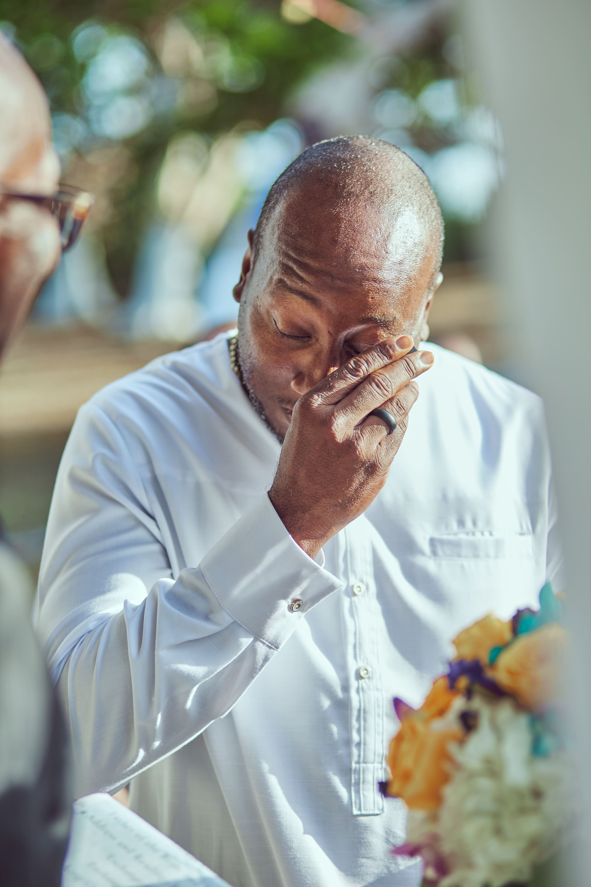 A man with a shaved head and dressed in a white button-up shirt is holding his face with his hand, appearing emotional or overwhelmed. There are blurred trees in the background and colorful flowers in the foreground.