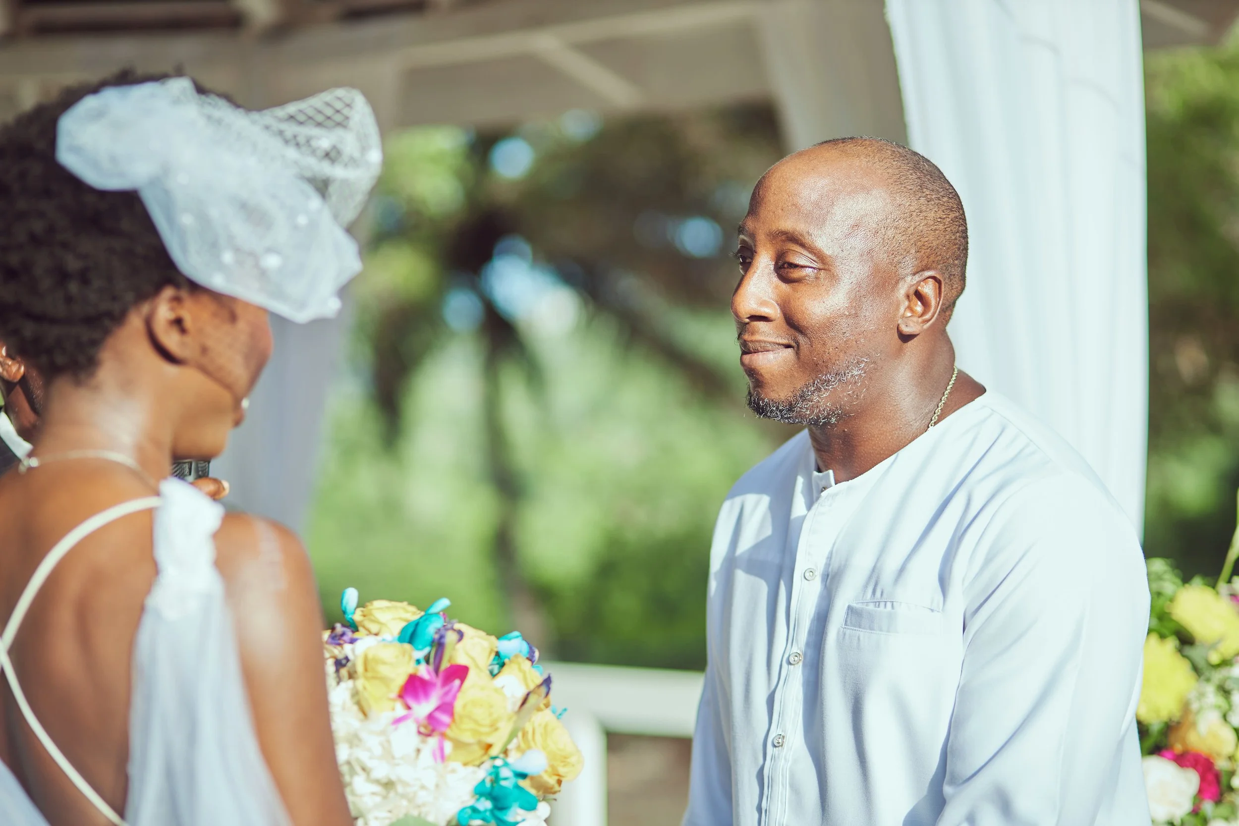 A man and woman exchanging vows during a wedding ceremony outdoors, with the woman holding a bouquet of yellow, purple, and pink flowers, and the man wearing a white shirt, in front of a background of green trees and white curtains.