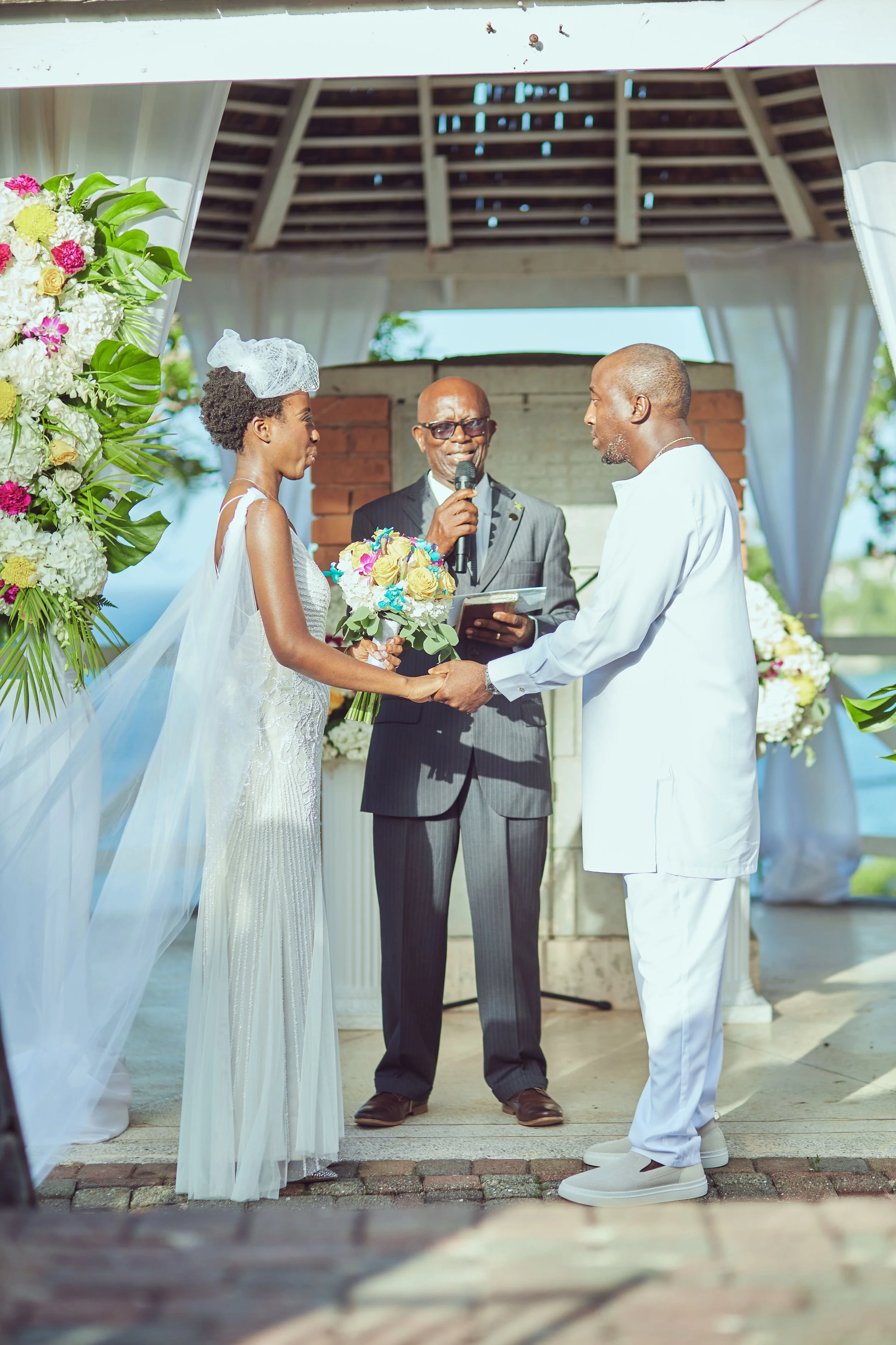 A couple exchanging wedding vows at an outdoor ceremony, with a minister officiating, in front of floral arrangements and a gazebo with ocean view in the background.