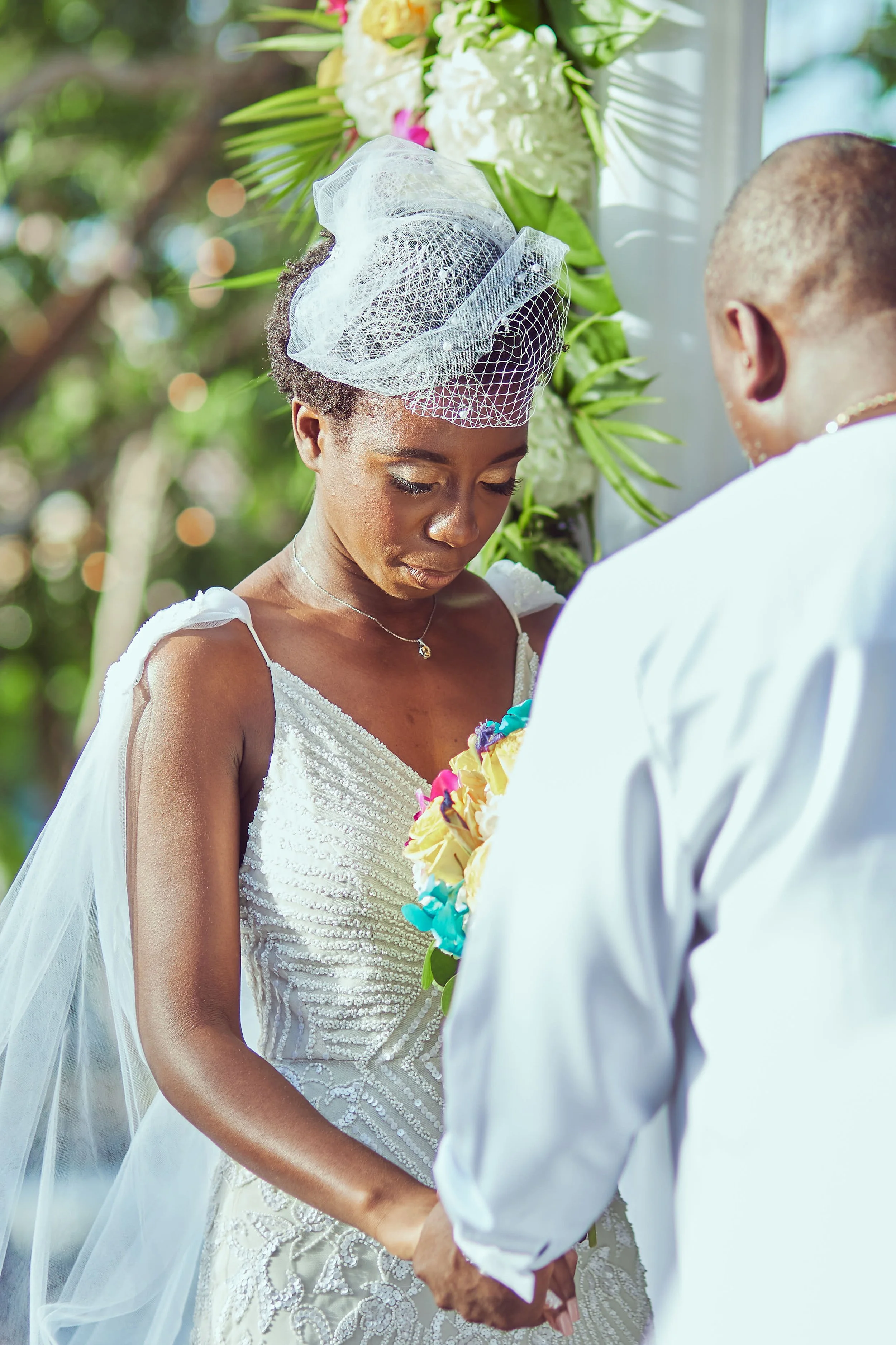 A bride and groom exchange vows outdoors during a wedding ceremony. The bride wears a white dress with intricate beadwork and a birdcage veil, holding a colorful bouquet. The groom, in a white shirt, faces her, and they hold hands in front of floral 