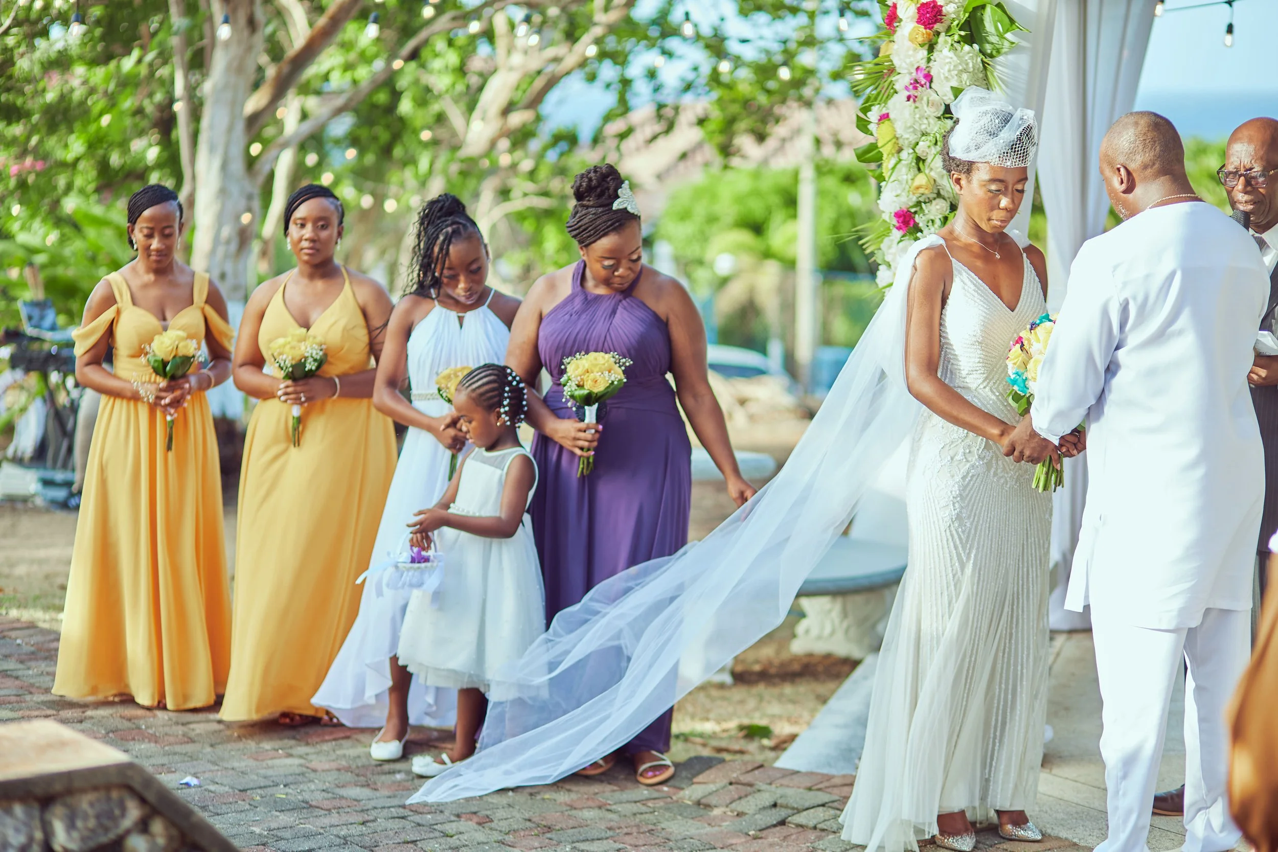 A wedding ceremony outdoors with the bride and groom exchanging vows, surrounded by bridesmaids and a flower girl holding bouquets, under a floral arch on a sunny day.
