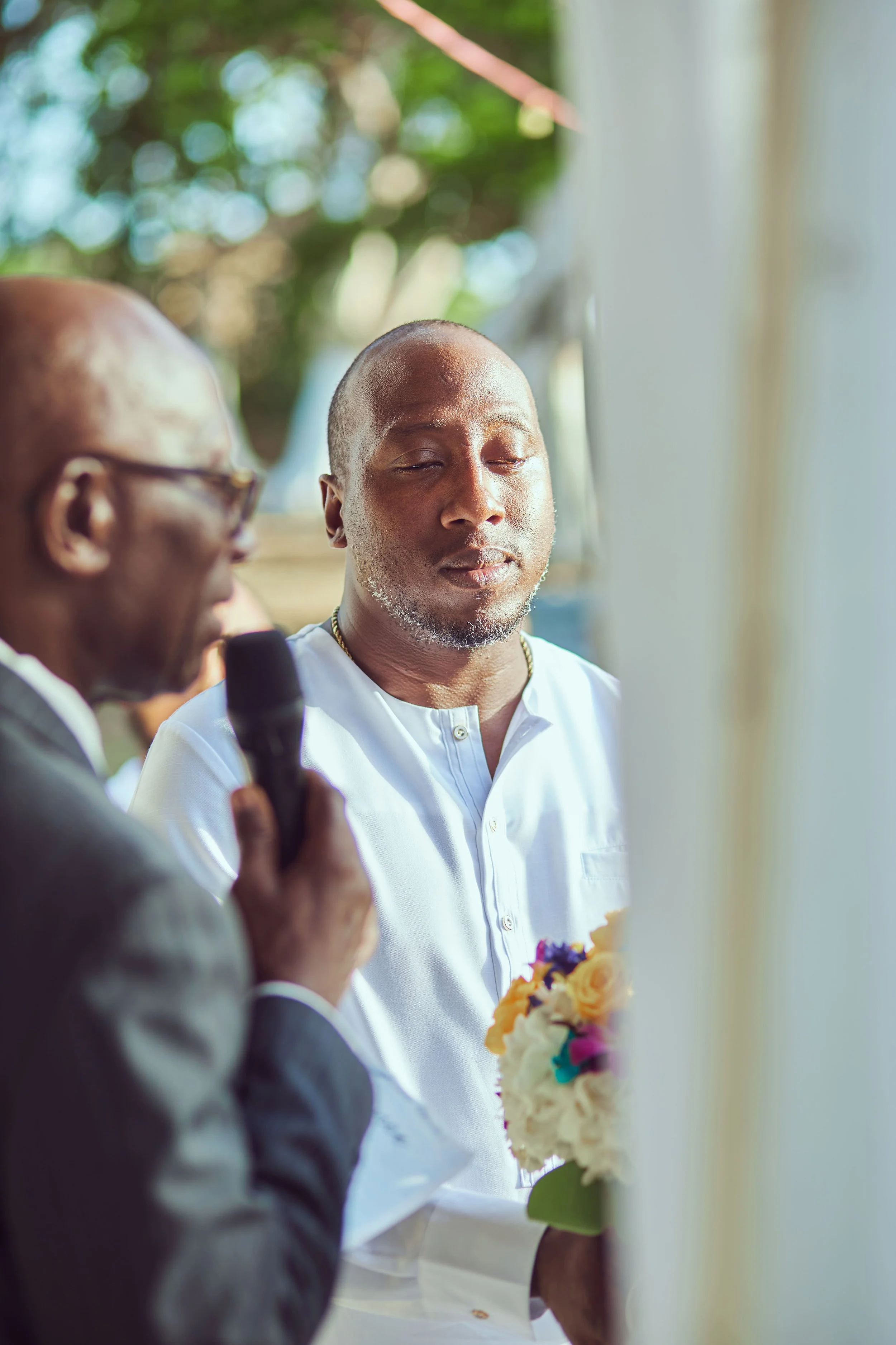 A man holding a bouquet of flowers and wearing a white shirt stands with his eyes closed during a ceremony. Another man, partially visible, holds a microphone and appears to be speaking. The background features blurry green trees and outdoor natural 