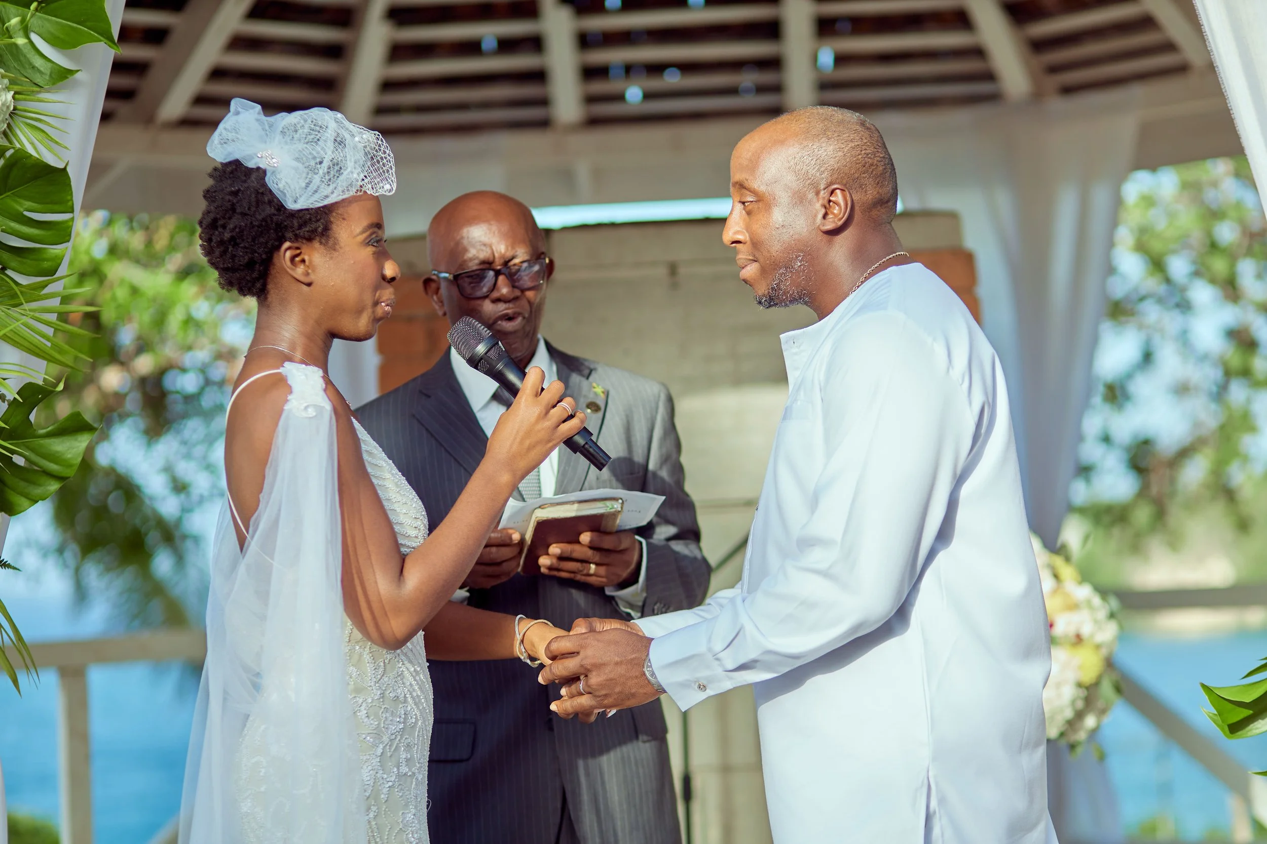 A couple exchanging vows during a wedding ceremony outdoors, with a officiant holding a microphone and a book, under a wooden pavilion decorated with flowers and greenery.