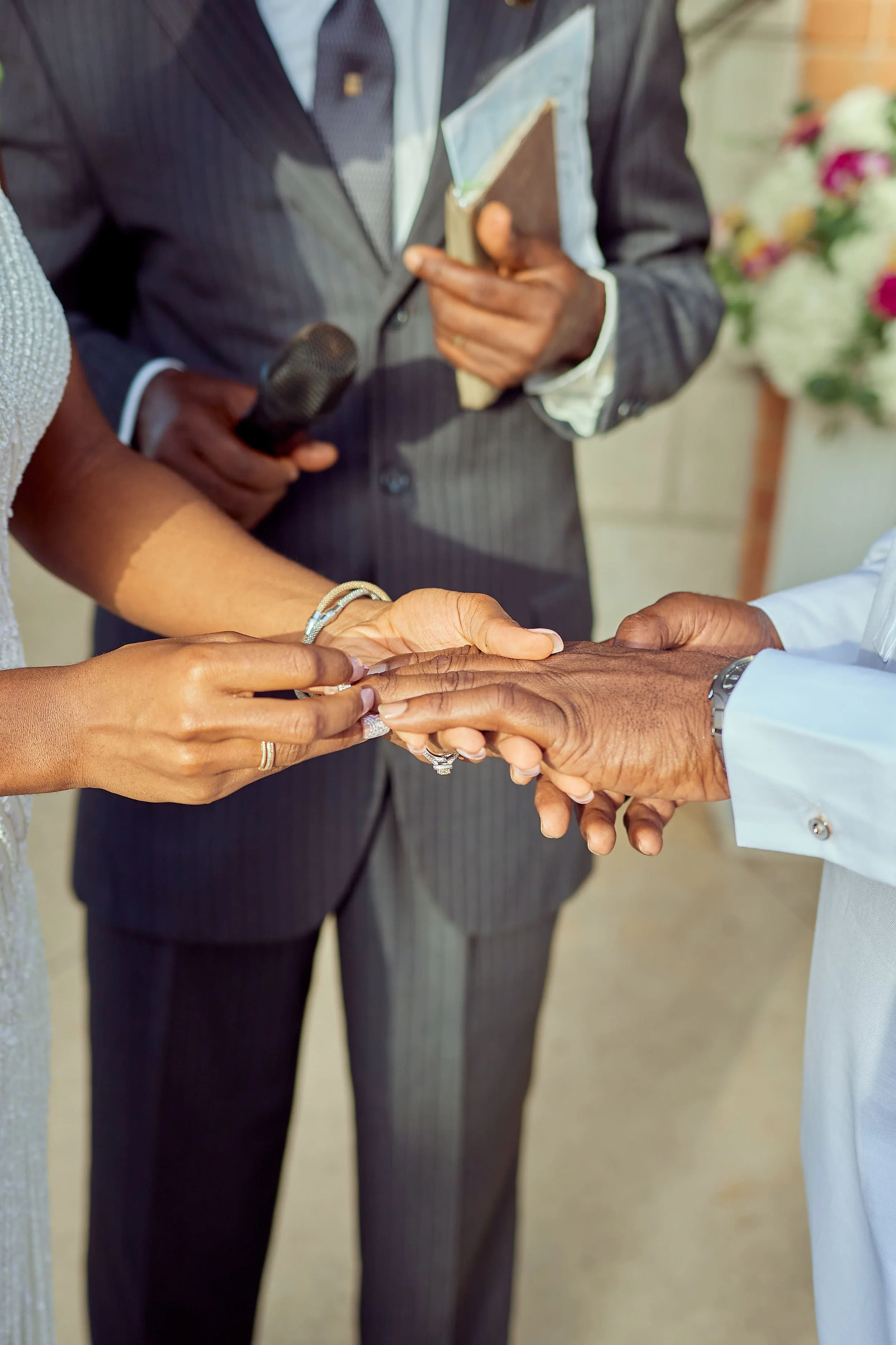 Close-up of a wedding ceremony with hands of the bride and groom exchanging rings, officiant in the background holding a microphone and a book, with floral decorations in the background.