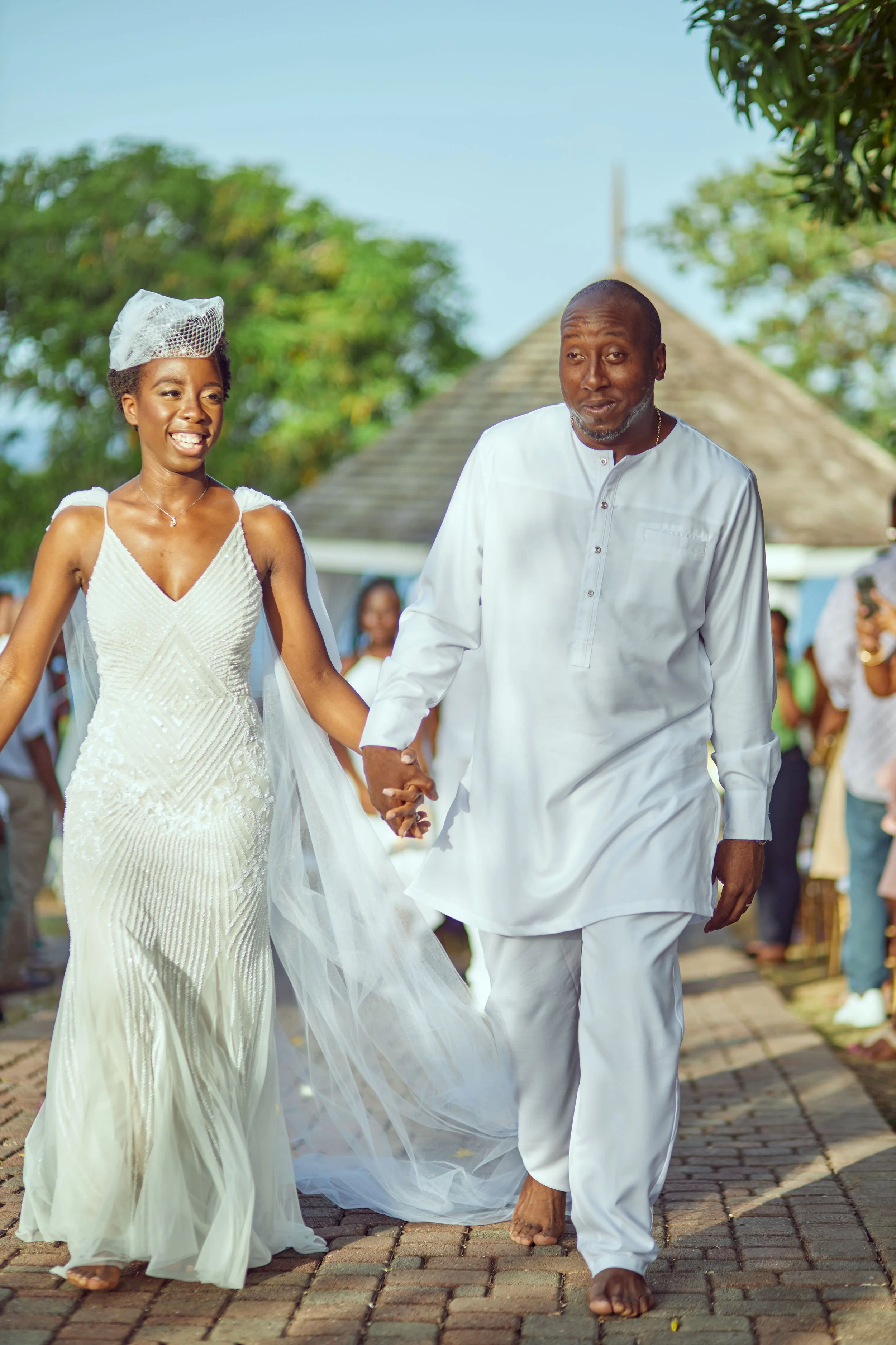 A bride and an older man, likely her father, holding hands and walking down the outdoor aisle during a wedding ceremony. The bride is smiling and wearing a white gown with a veil, and the man is dressed in traditional white attire. Guests are visible