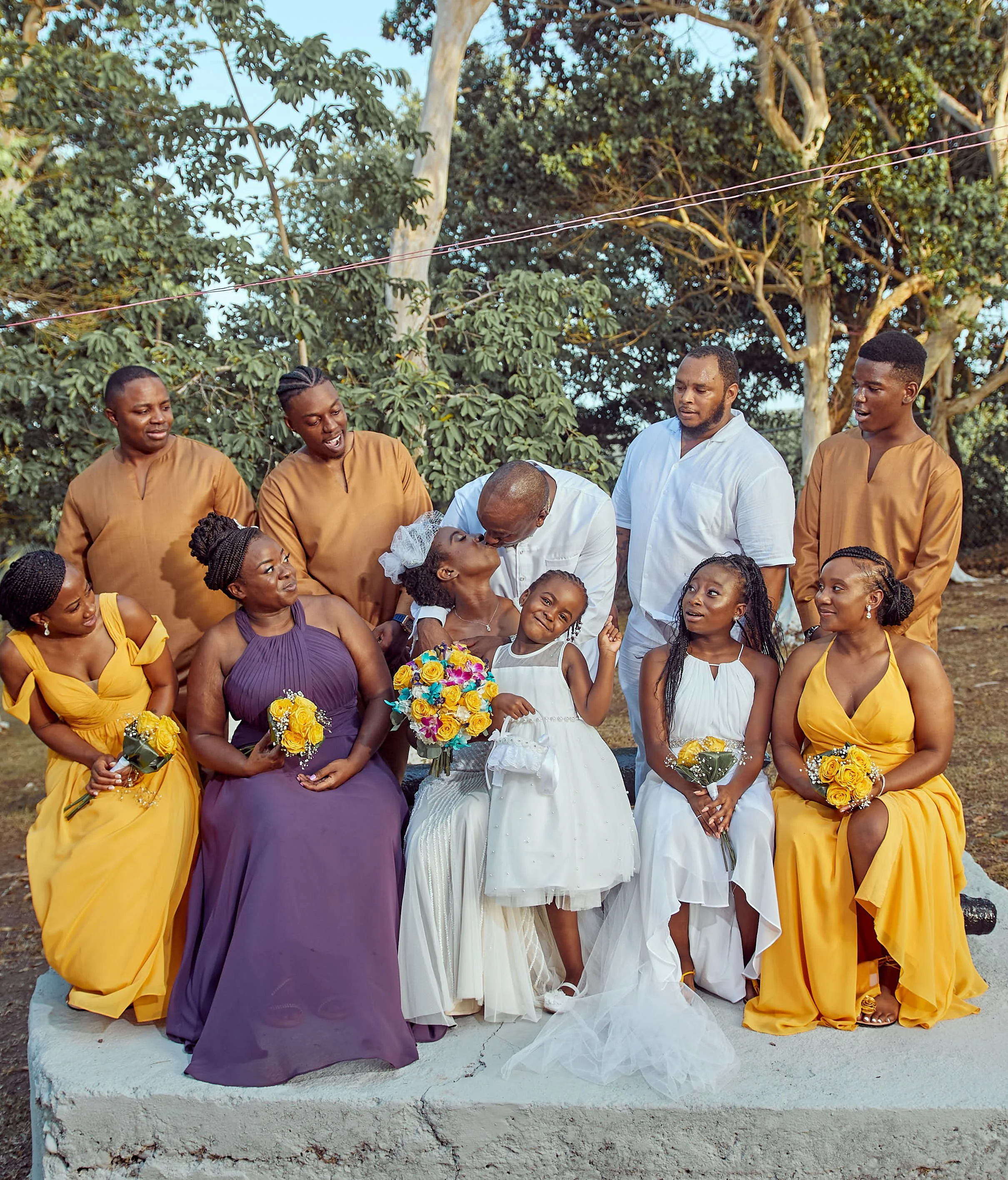 A group of people gathered outdoors for a celebration, possibly a wedding, with greenery and trees in the background. Some are holding bouquets of flowers and two children are present in the center.
