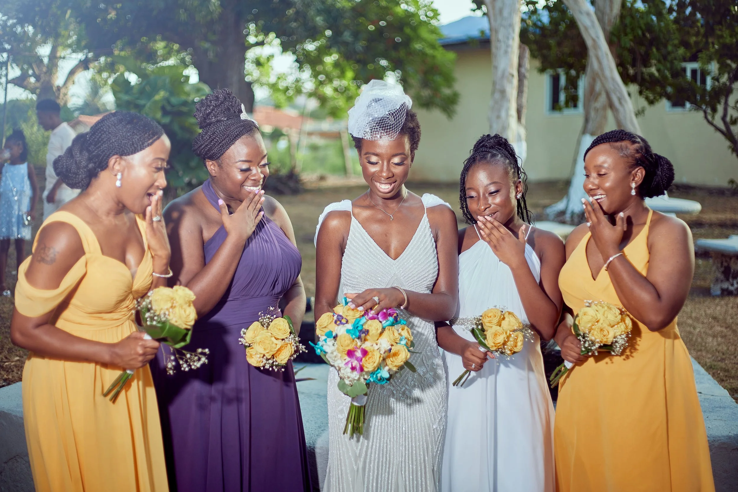 A group of women dressed in colorful dresses, standing outdoors during a wedding celebration, smiling and holding bouquets of yellow roses.