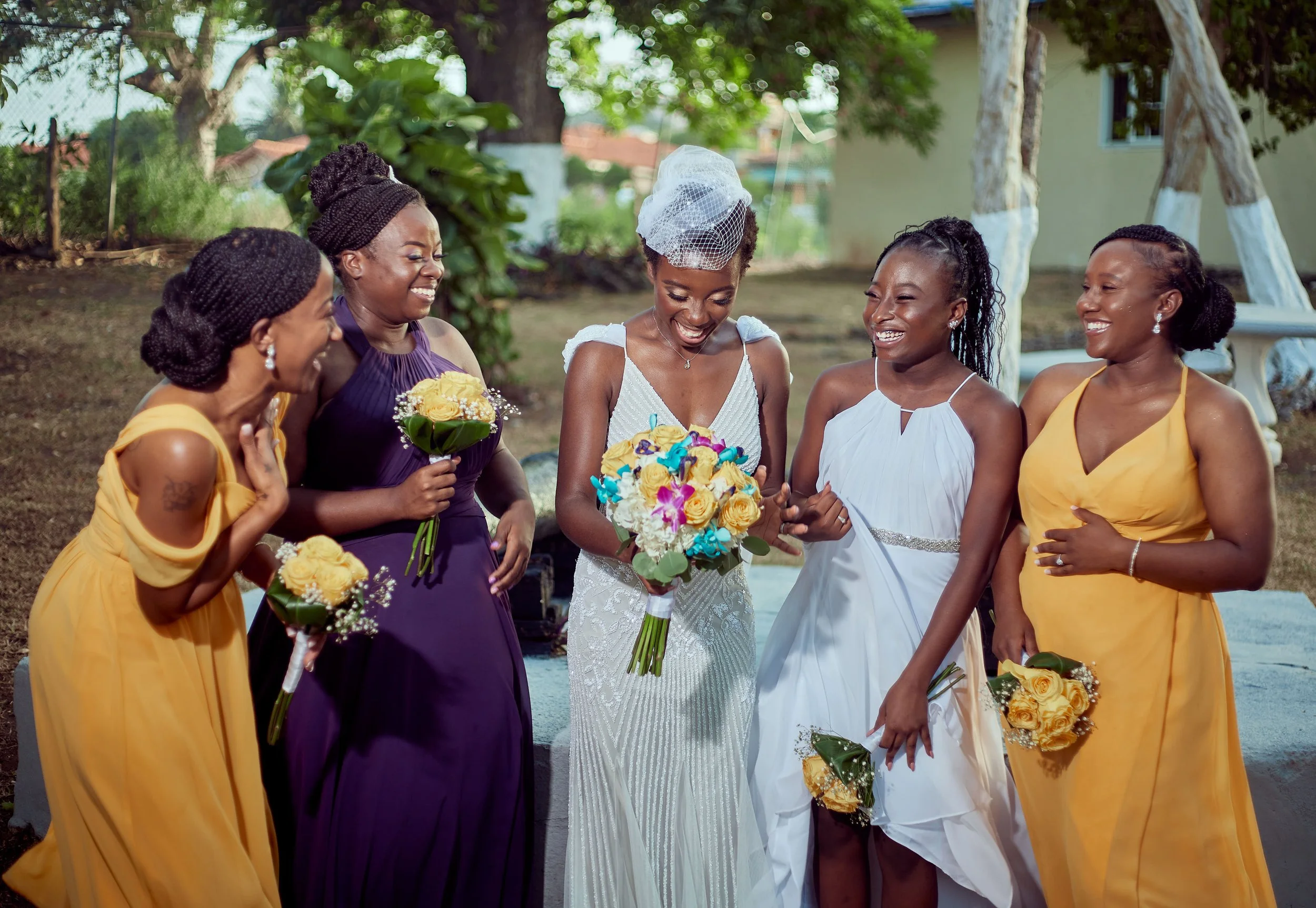 A bride in a white wedding dress holding a bouquet of flowers, surrounded by five women in colorful dresses, outdoors with trees and a building in the background.