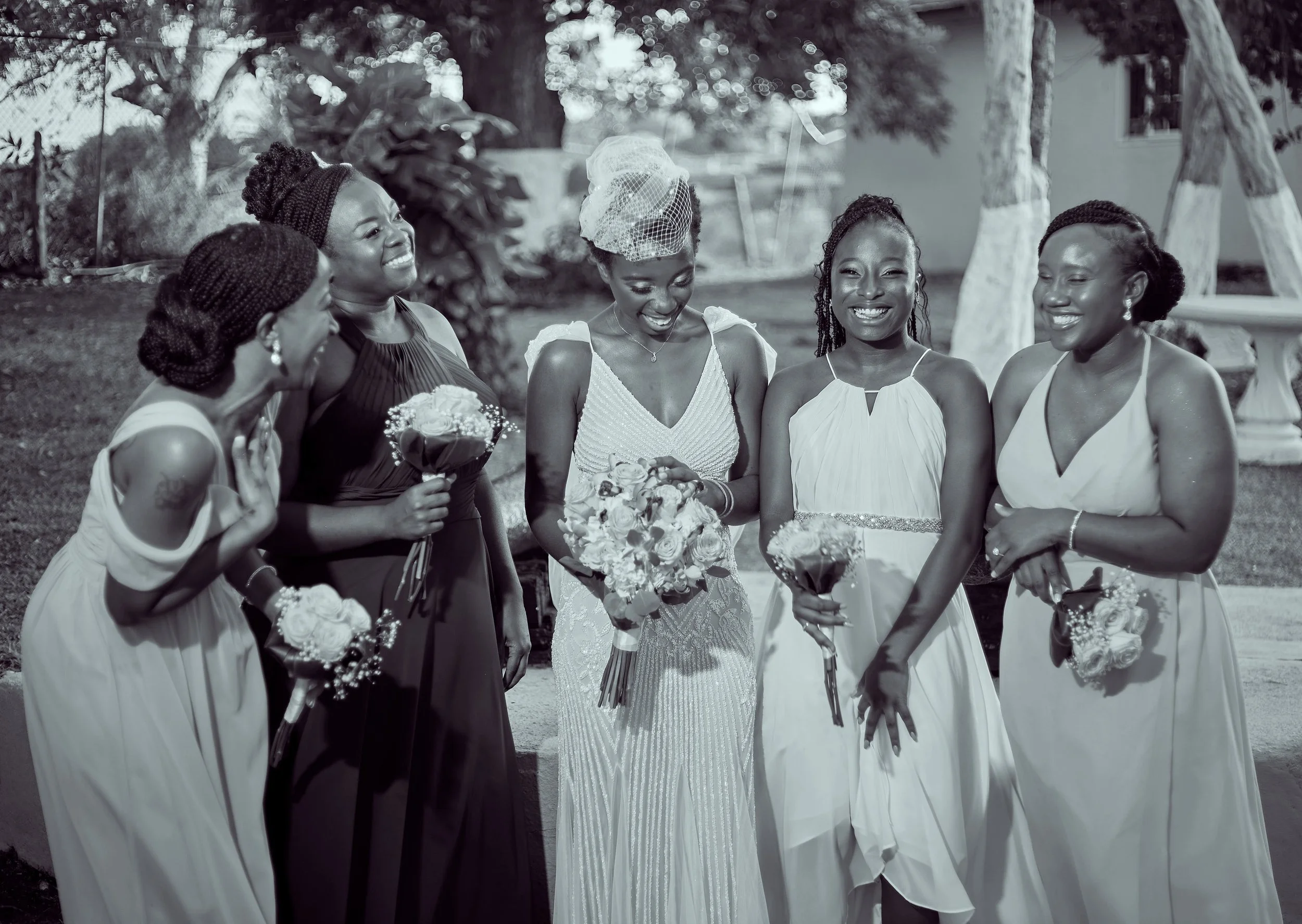 Five women in formal dresses standing outdoors, smiling and holding bouquets, celebrating a wedding.