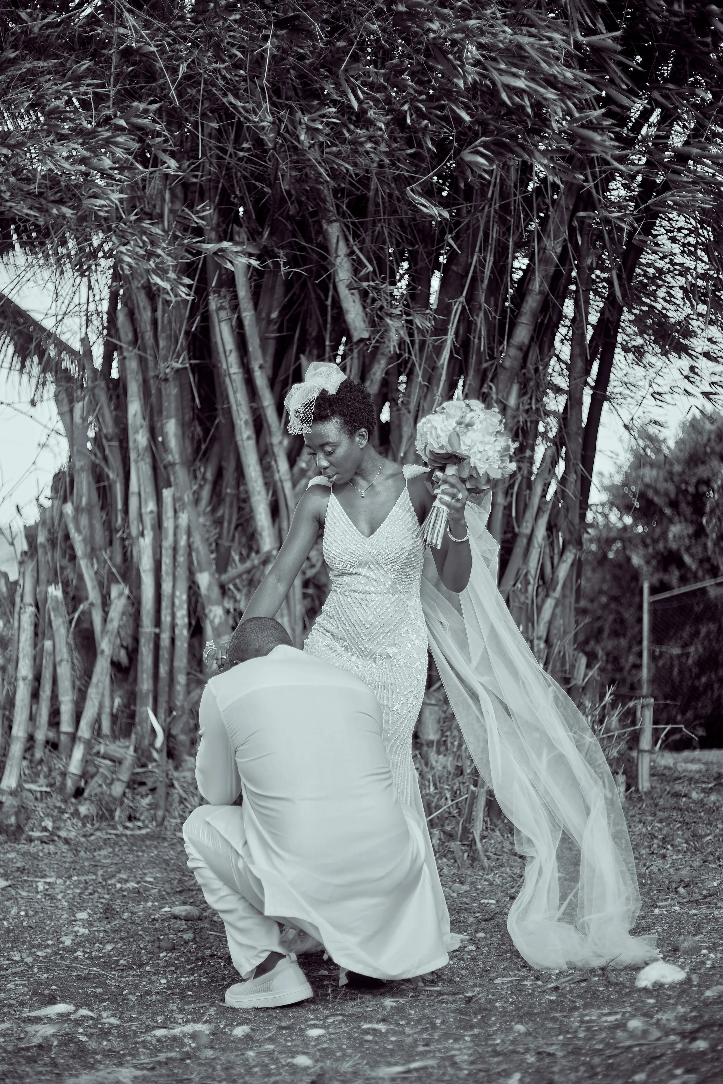 A woman in a wedding dress holding a bouquet, standing outdoors in front of tall bamboo plants, with a man kneeling before her.