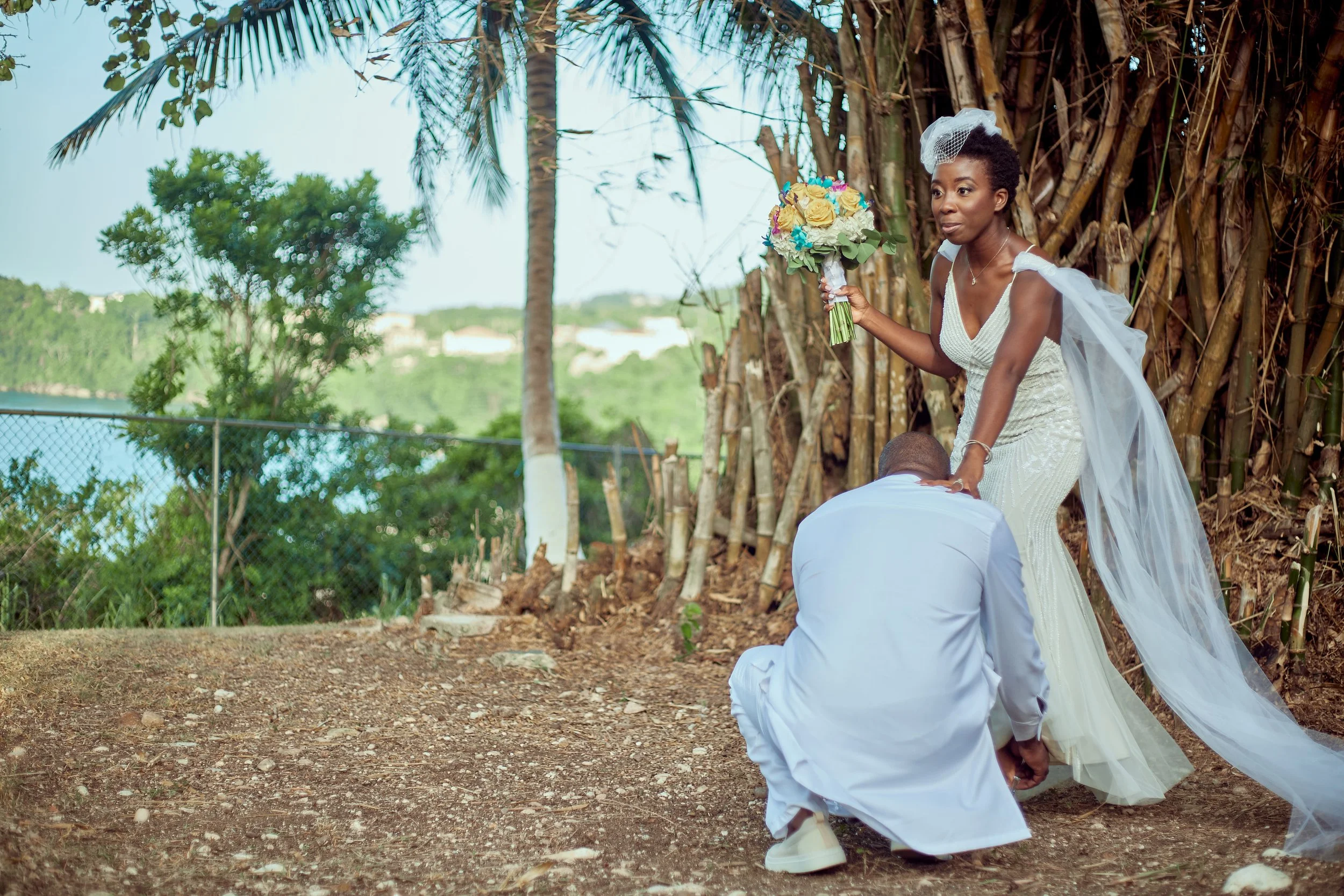 A couple getting married outdoors near a large tree, with the bride holding a bouquet of flowers and the groom kneeling in front of her.