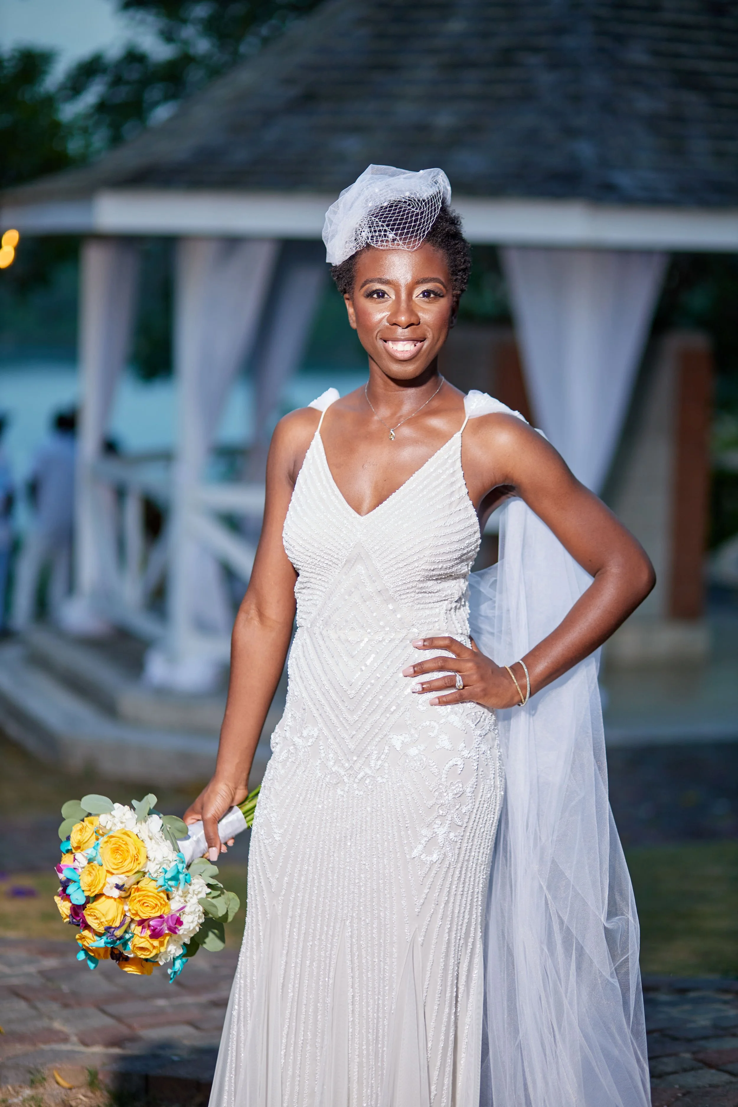 A woman in a white wedding dress holding a colorful bouquet, standing outdoors with a pavilion in the background.