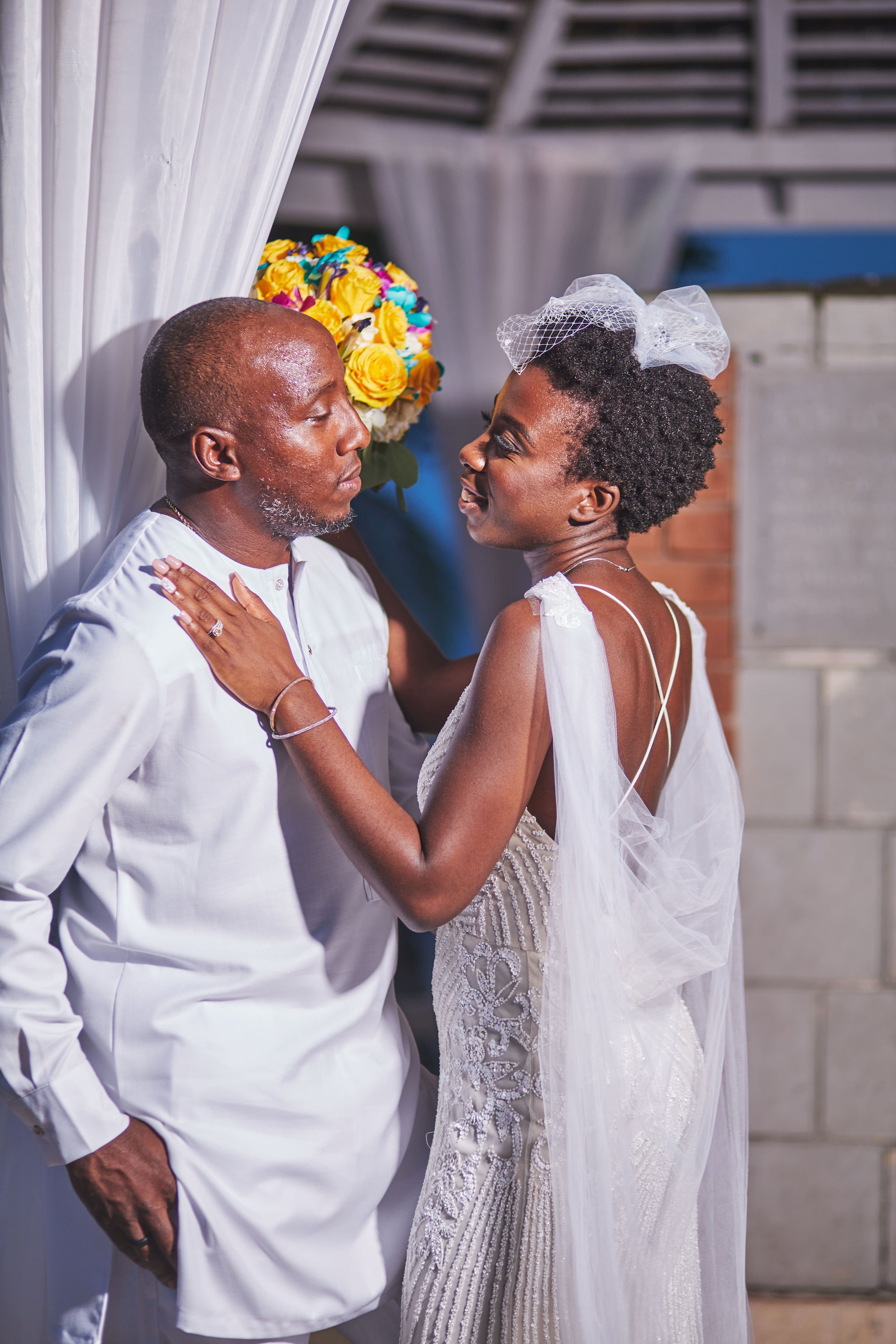 A couple dressed in wedding attire standing close together, with the woman reaching out to touch the man's chest, in front of a colorful bouquet of yellow and blue flowers.