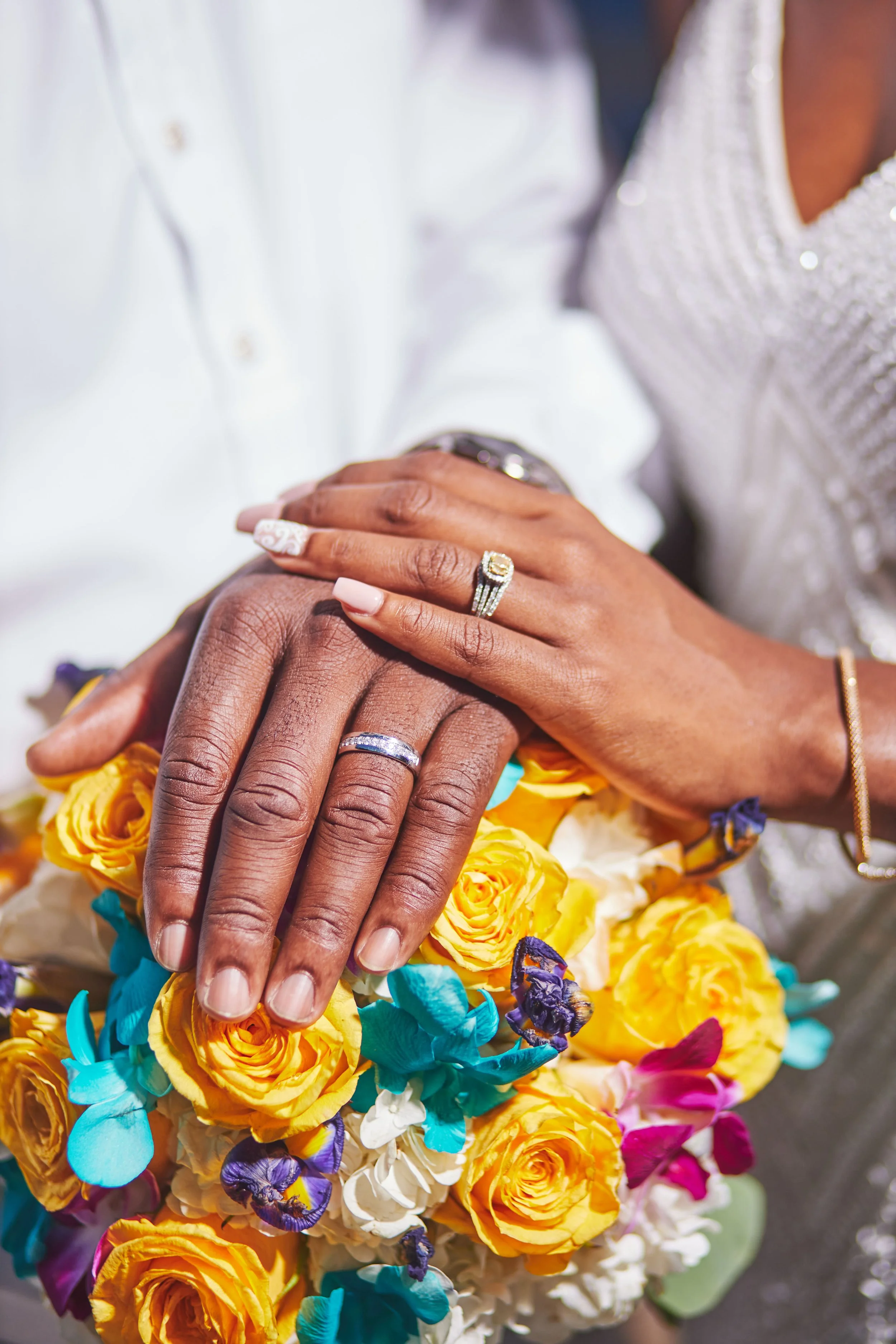 Close-up of a couple's hands showing wedding rings, resting on a bouquet of colorful yellow, white, purple, pink, and teal flowers.