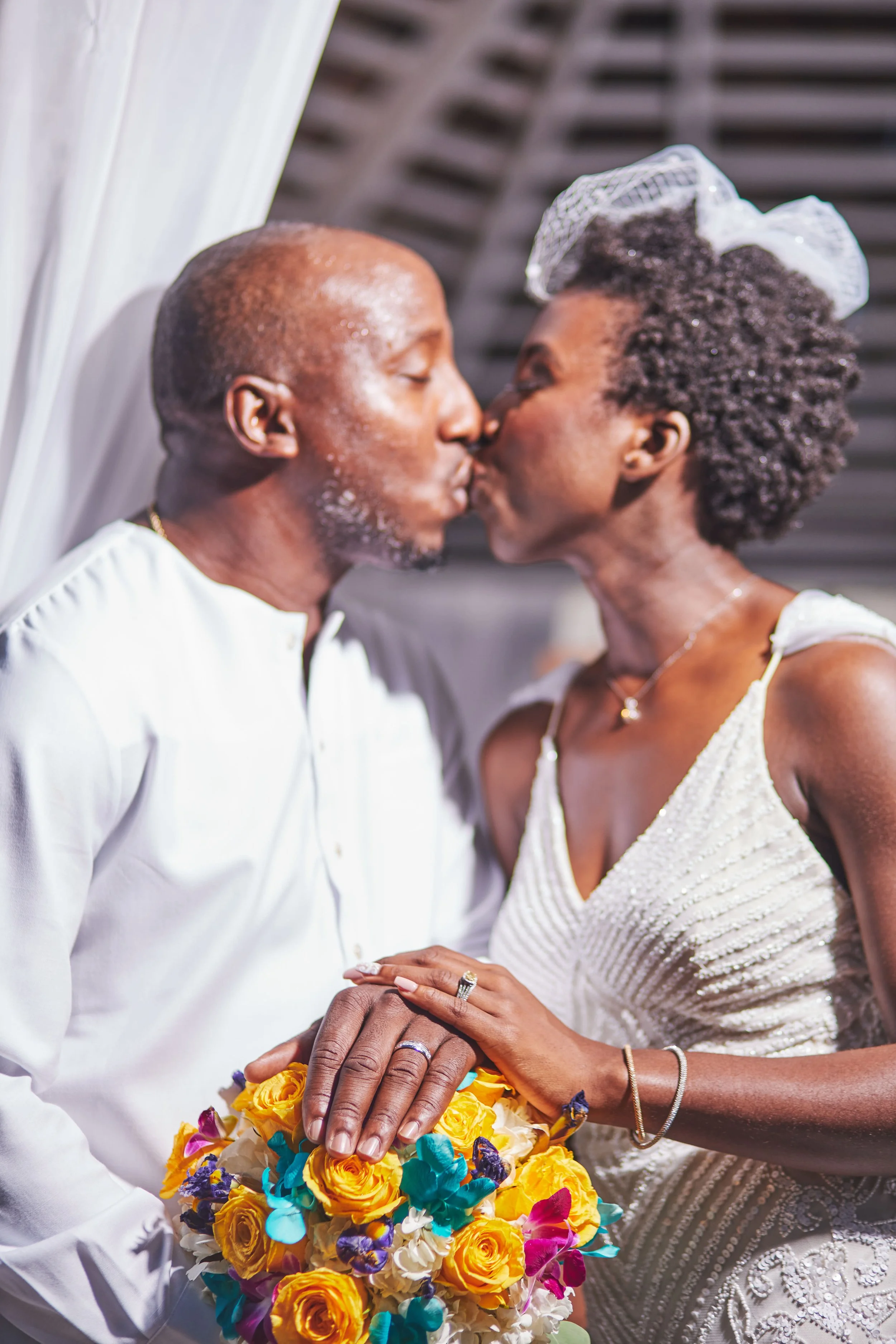 An elderly couple sharing a kiss at their wedding, holding a bouquet of yellow and purple flowers, with both wearing wedding rings and elegant white attire.