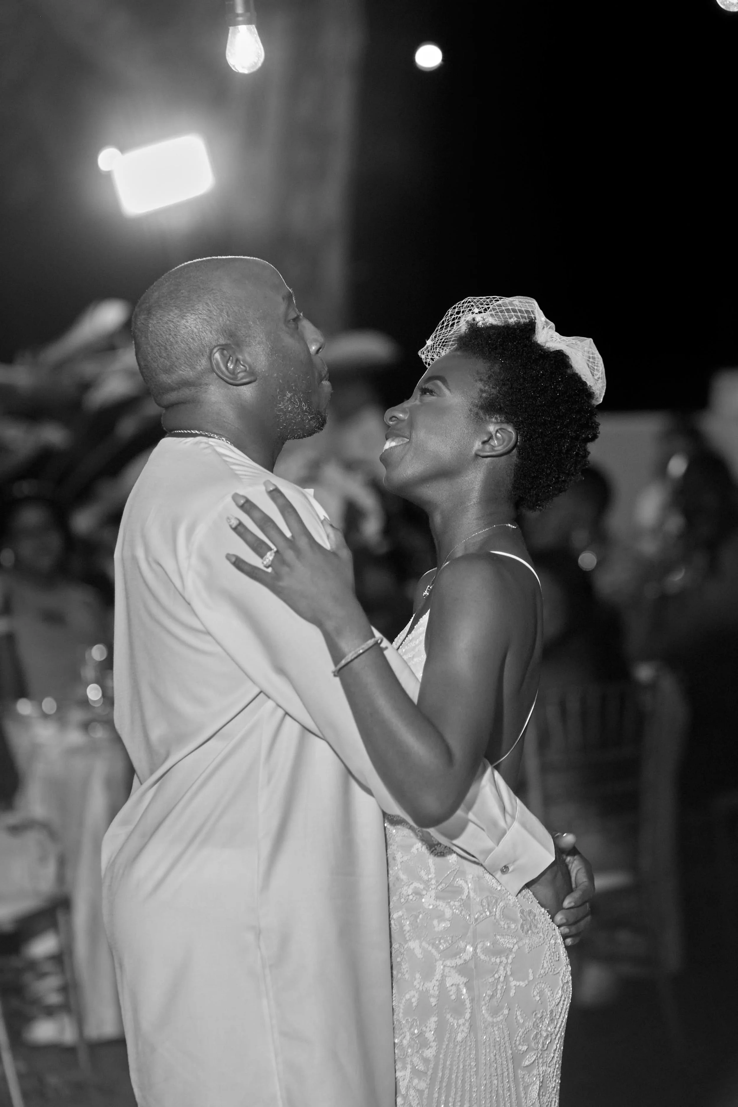 A couple dancing closely at a wedding reception, with the woman wearing a lace dress and a small veil, and both looking at each other lovingly, in a black-and-white photo.