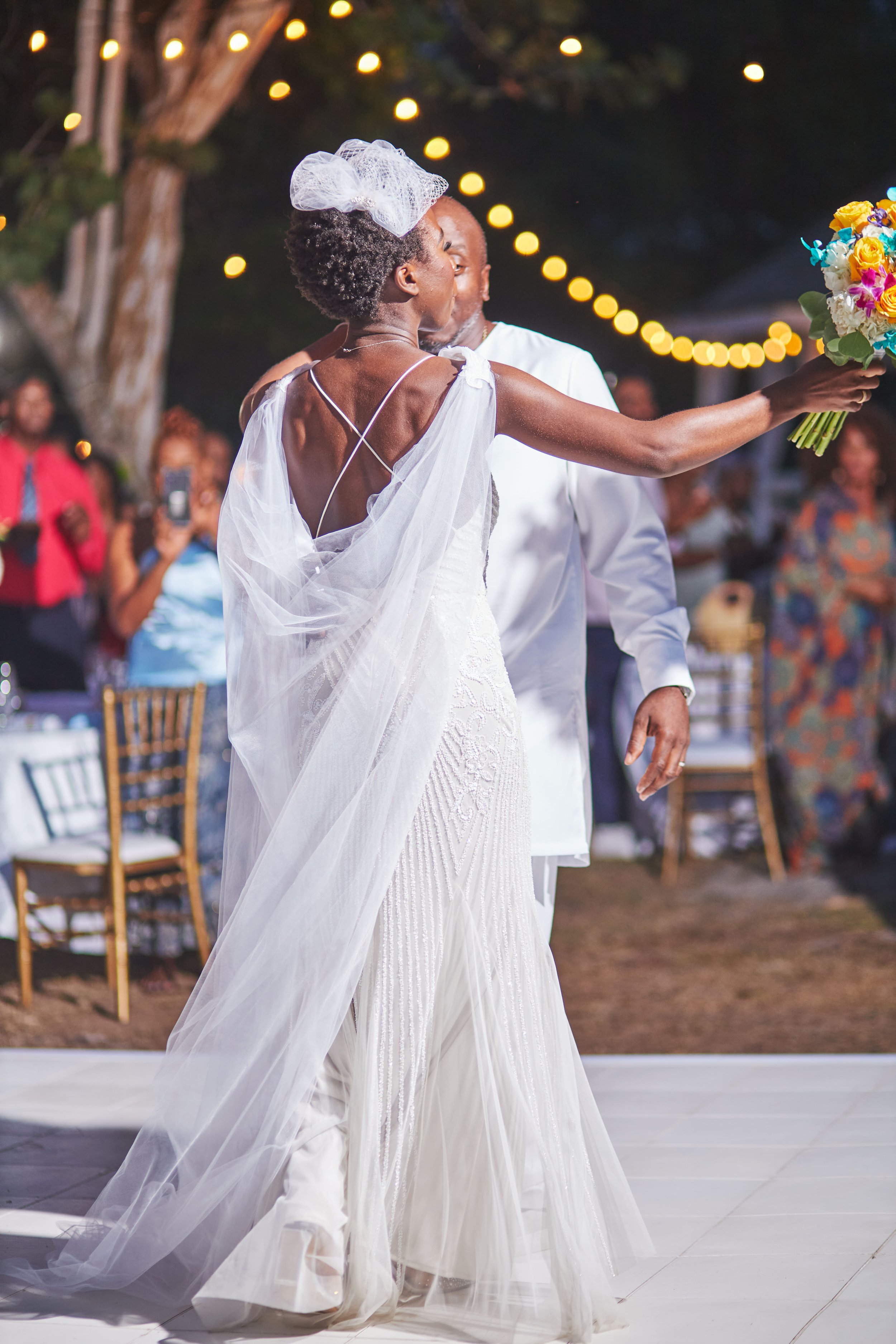 A bride and groom dancing at their outdoor wedding reception during evening, with string lights overhead and guests in the background.