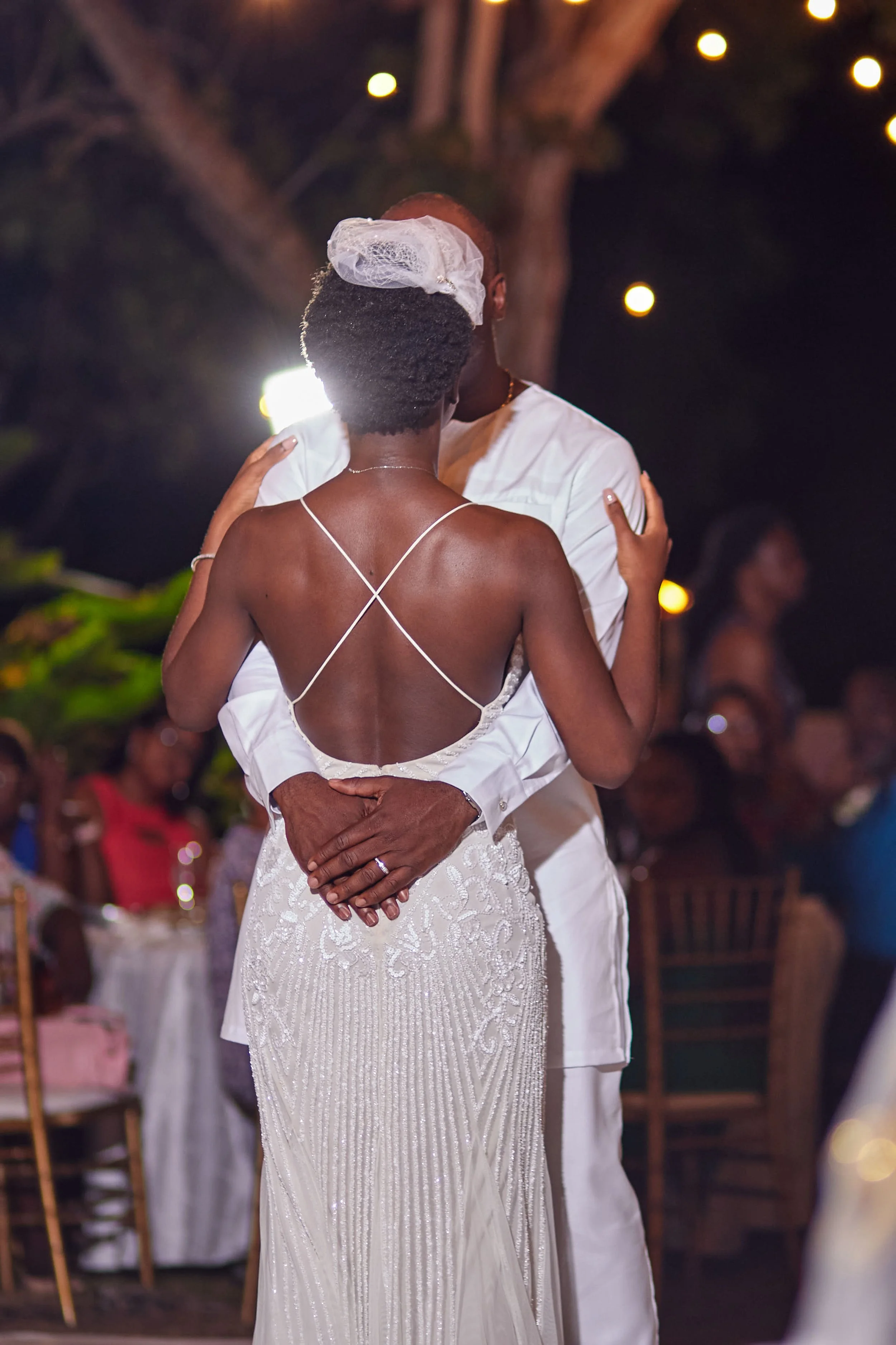 A couple dancing closely at their wedding reception during the evening, with guests seated at tables in the background and string lights overhead.