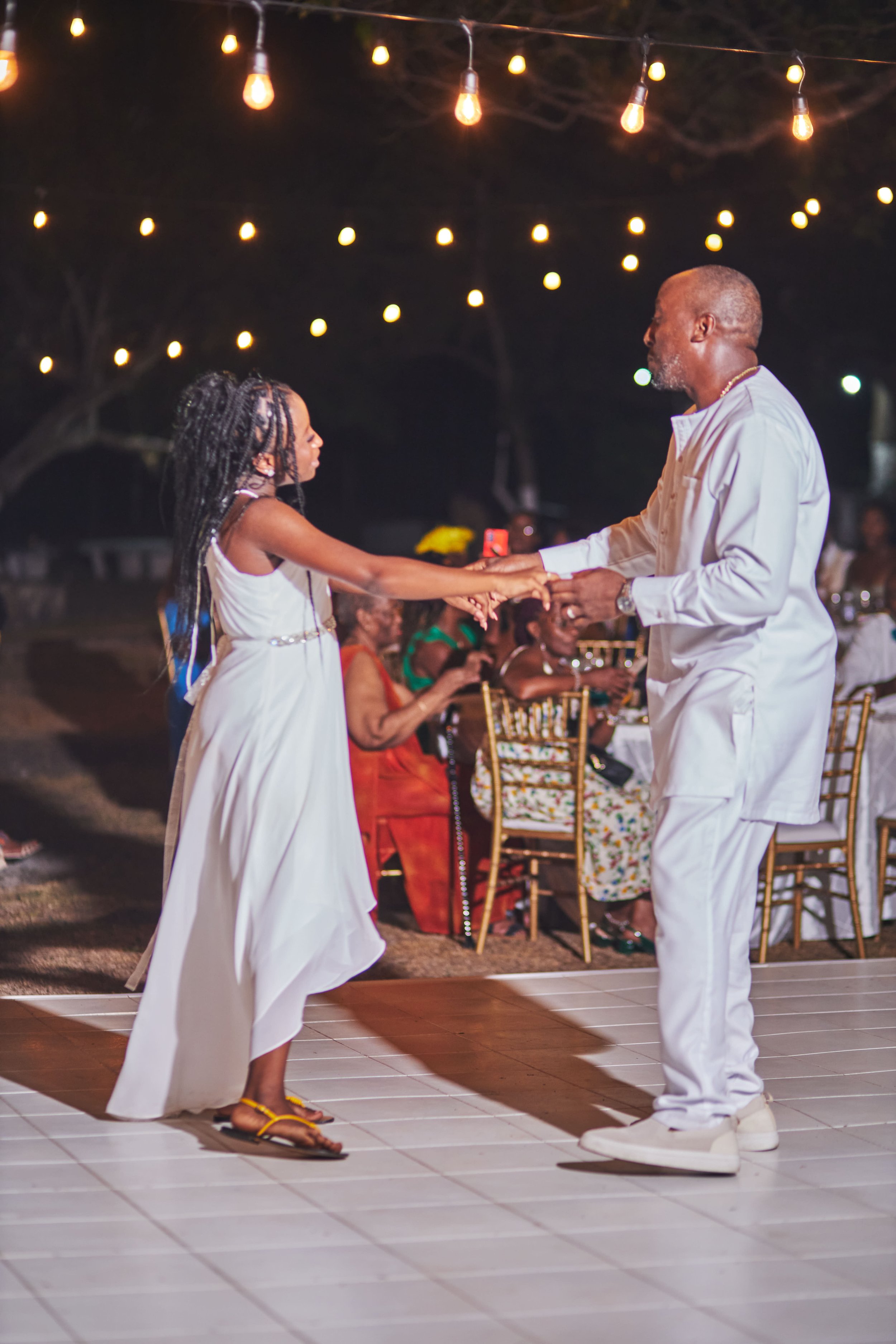 A girl and man dancing together at an evening outdoor event with string lights overhead, surrounded by seated guests.
