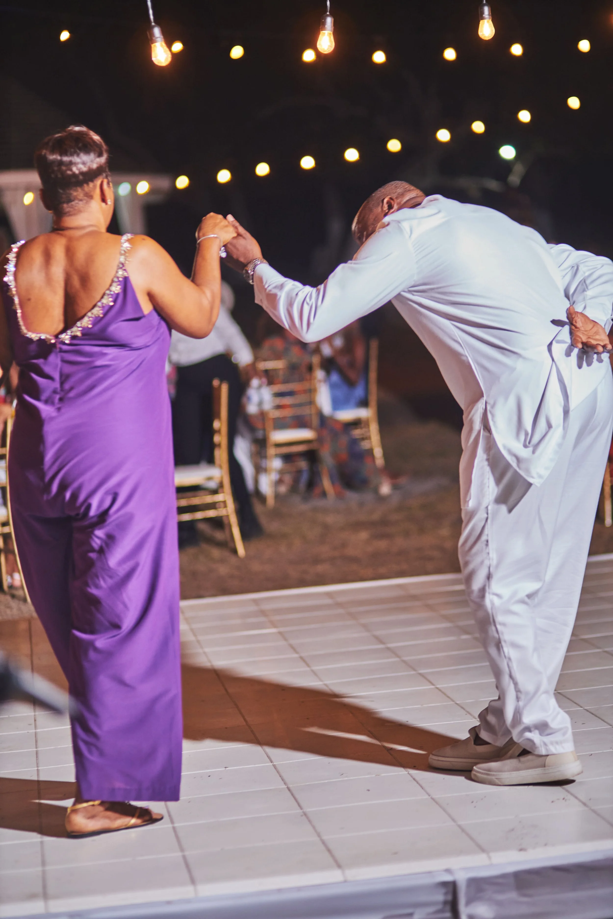 A man and a woman dancing together at an outdoor event at night, with string lights overhead and chairs in the background.