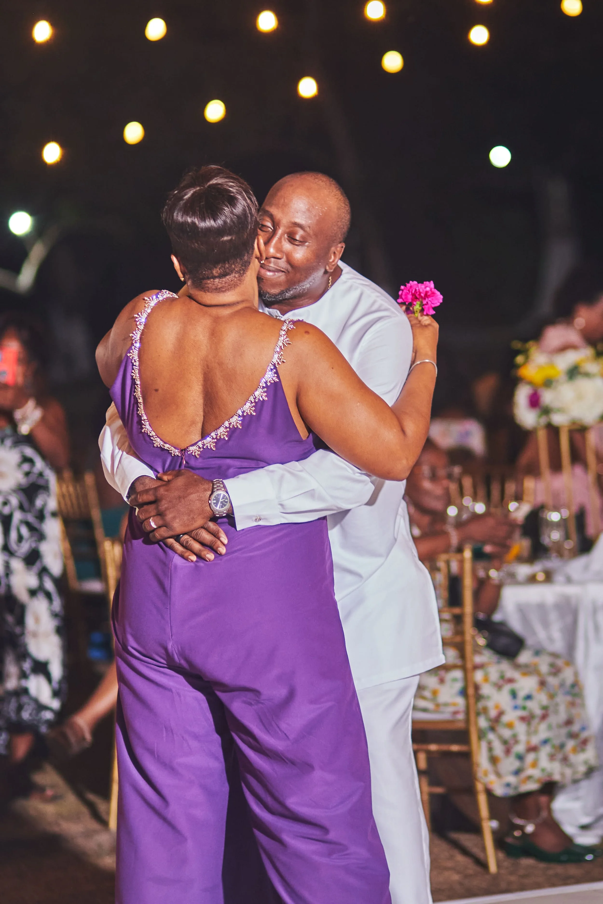 A couple dancing closely at a celebration, with the woman holding a pink flower and the man wearing a white shirt. Guests are seated at tables with floral centerpieces in the background.