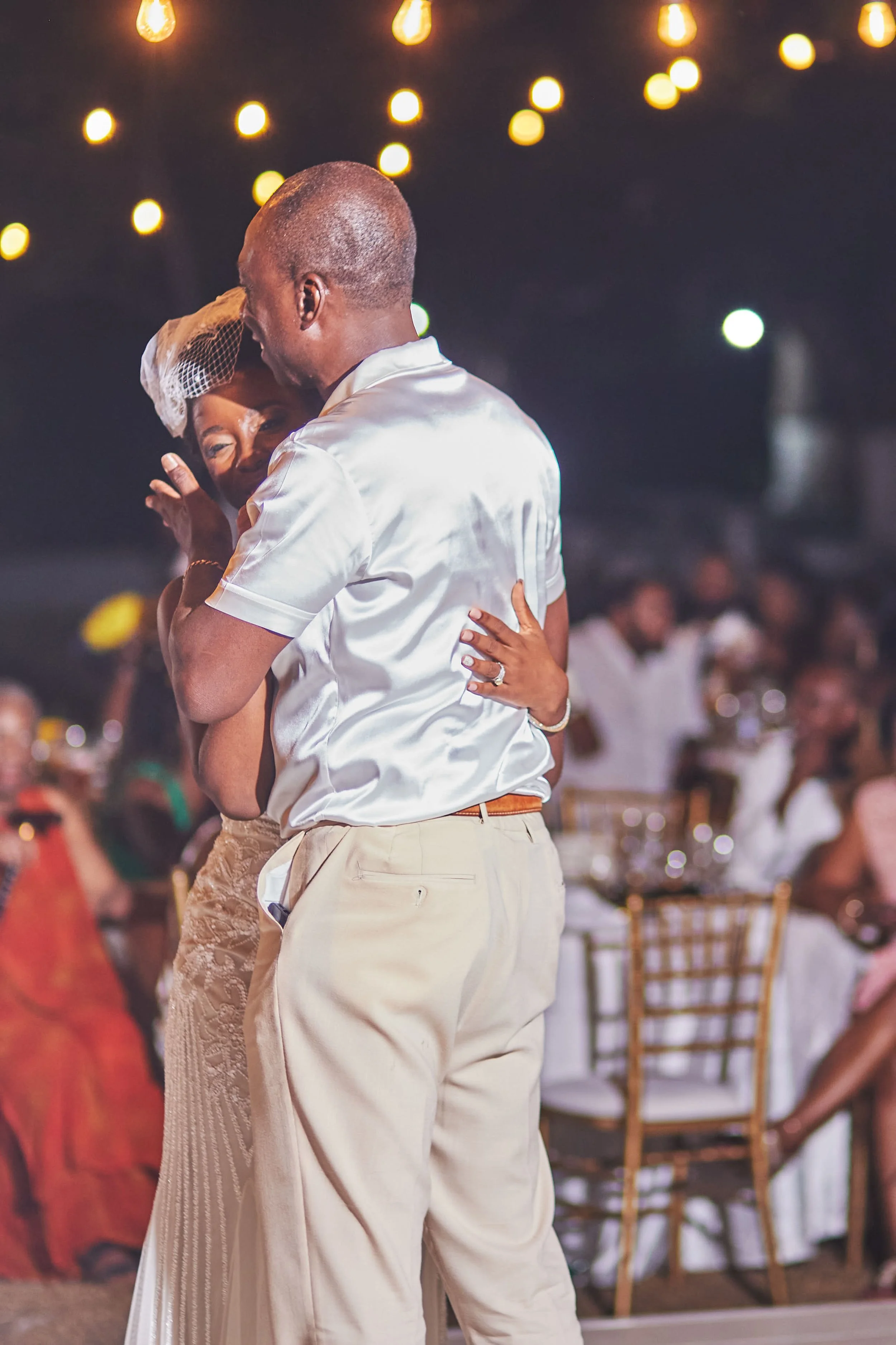 A close-up of an older man and woman dancing together at a wedding reception, with warm string lights in the background.