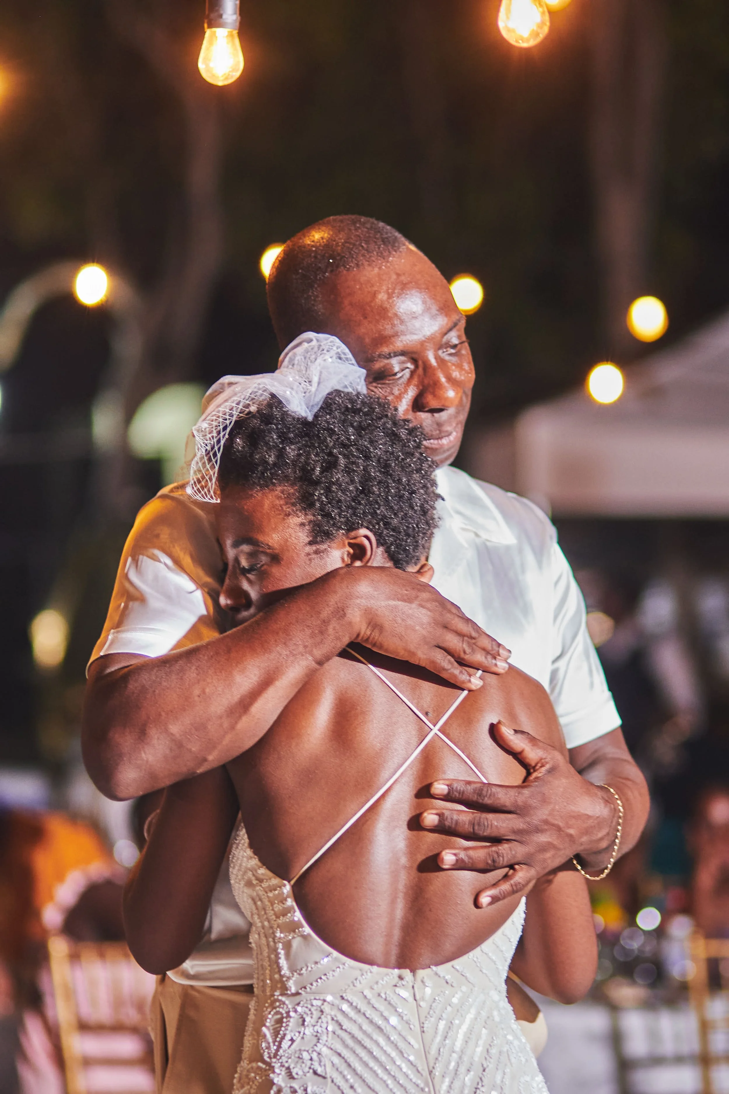 A man and a young girl share an emotional hug at a celebration or wedding, with warm hanging lights in the background.