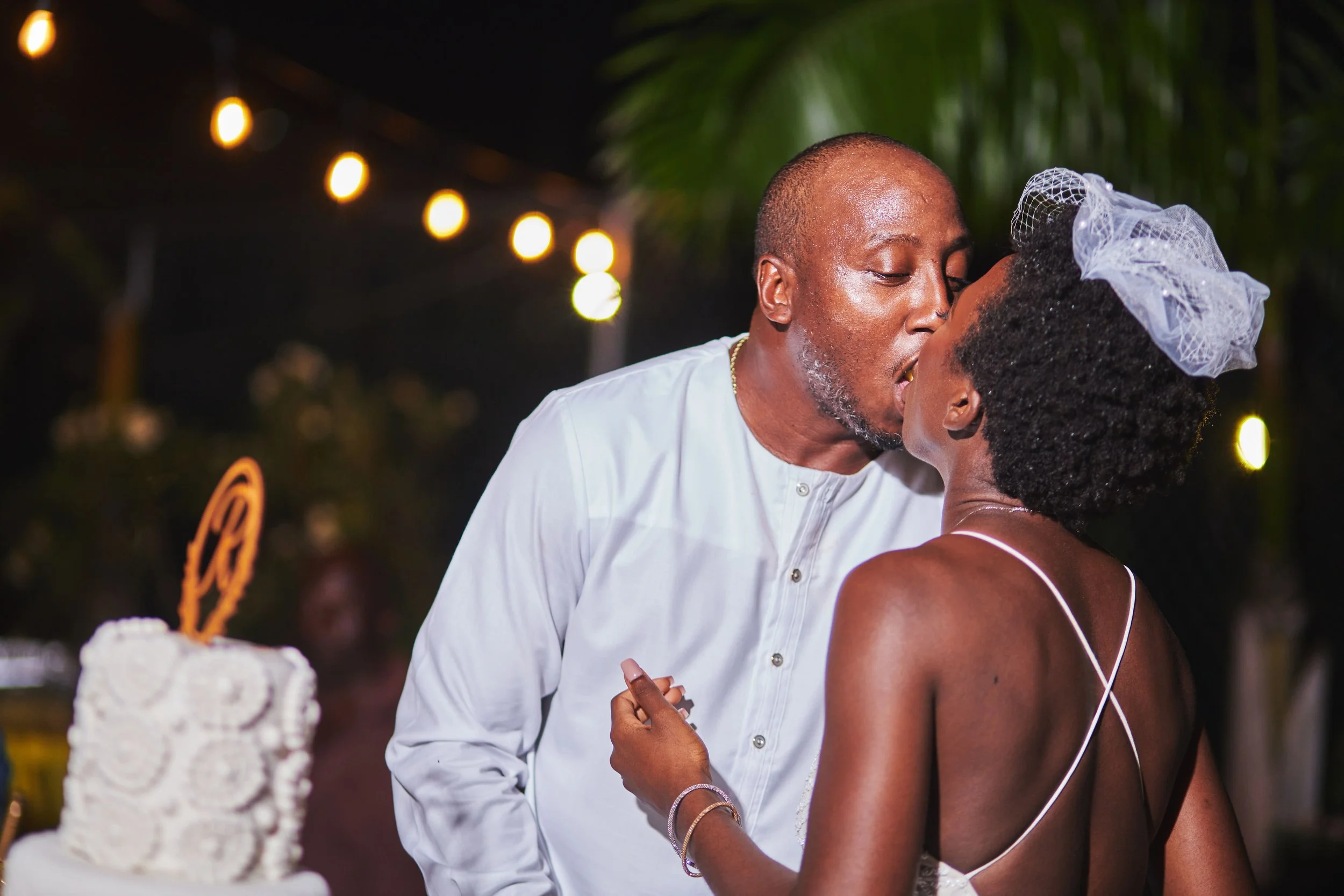A couple sharing a kiss during a wedding celebration at night, with string lights in the background.