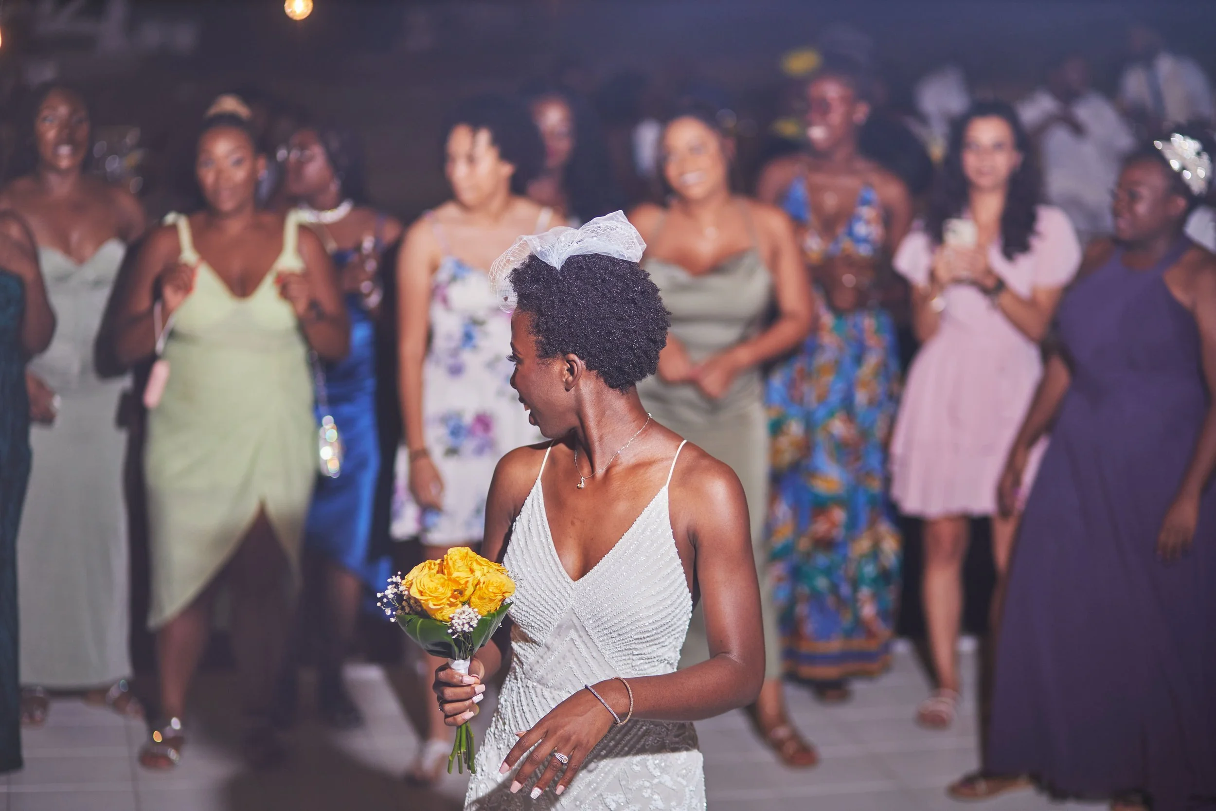 A woman in a white dress holding a bouquet of yellow flowers, with a veil on her head, is facing sideways. She is at a celebration or party with other women dancing in the background.