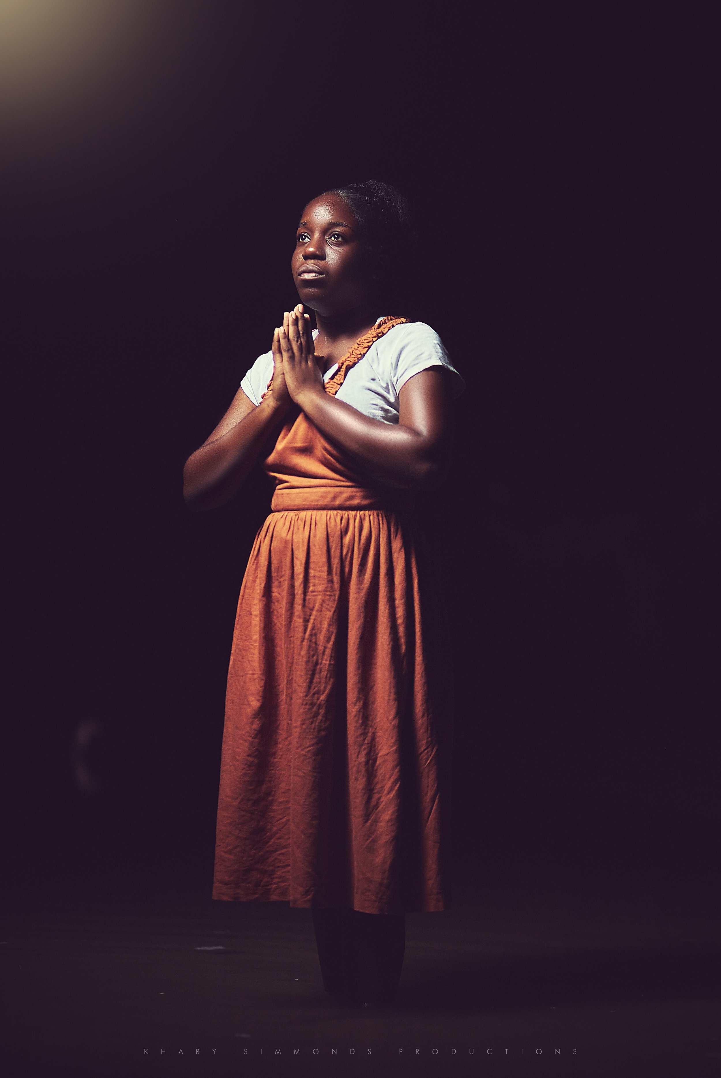 A young girl with dark skin standing with her hands together in prayer or meditation, wearing an orange skirt and a white T-shirt in a dimly lit setting.