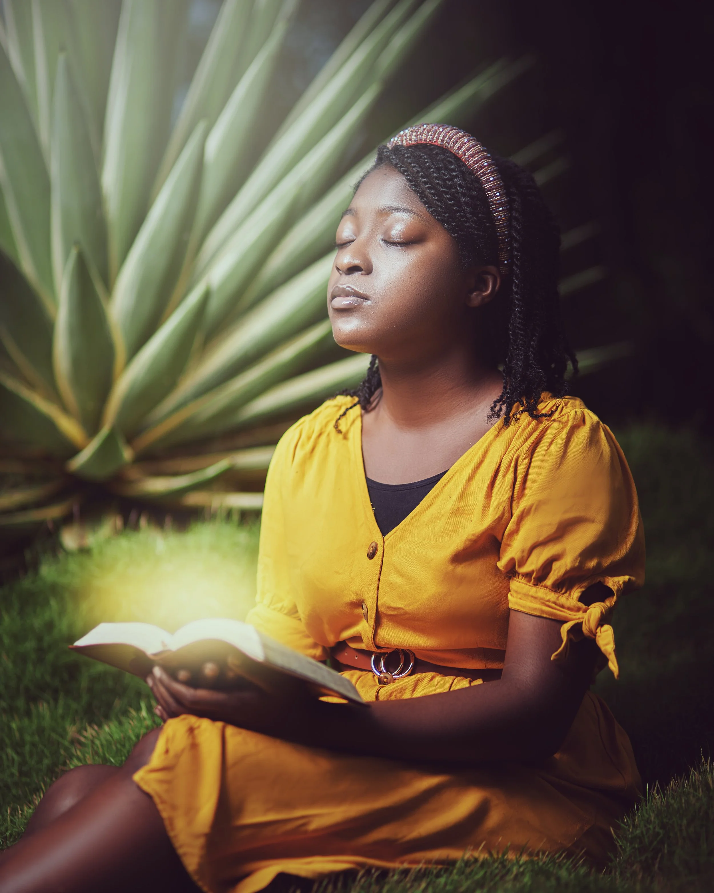A woman sitting outdoors with eyes closed, holding an open book emitting a glowing light, with a large agave plant in the background.