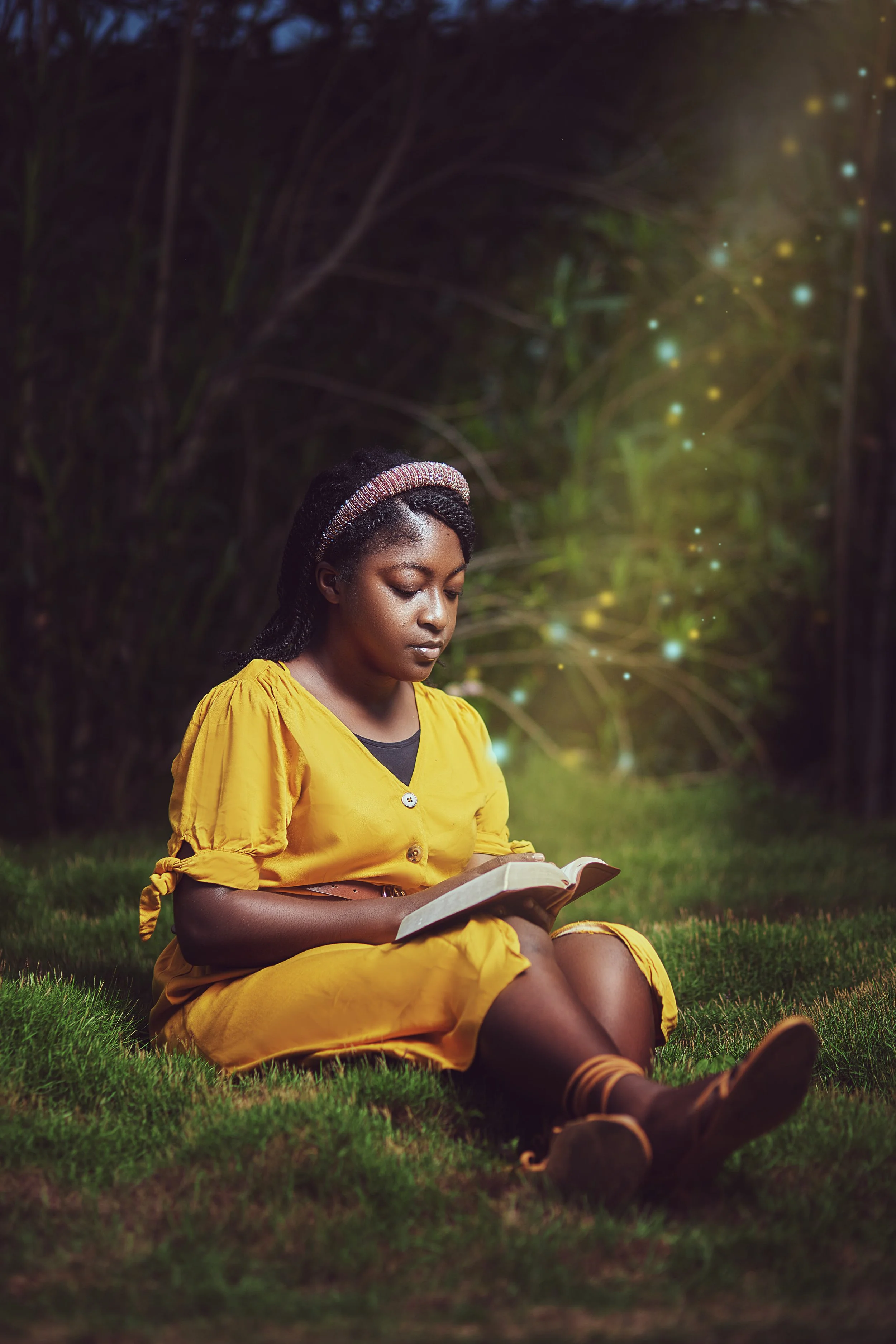 A young girl sitting on grass outdoors, reading a book with a dark background and glowing light or sparkles near her, wearing a yellow dress and a headband.