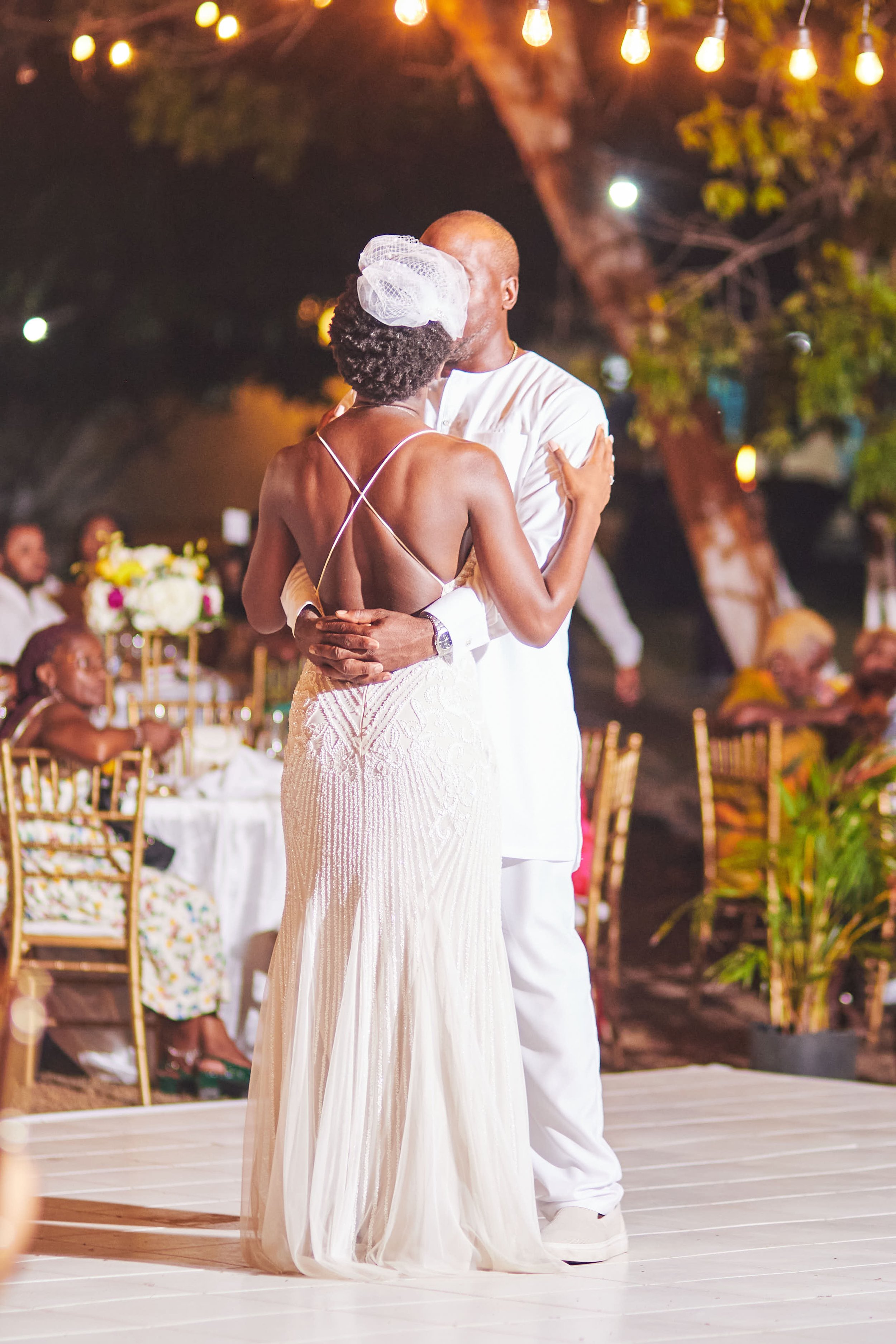A couple dancing closely at an outdoor wedding reception, with guests seated at tables in the background and string lights overhead.