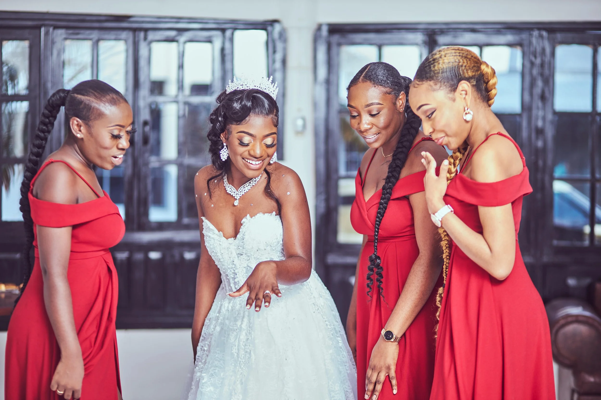 A bride in a white wedding dress and tiara is smiling and showing her ring, surrounded by four women in red dresses, all smiling and looking at the bride's hand.